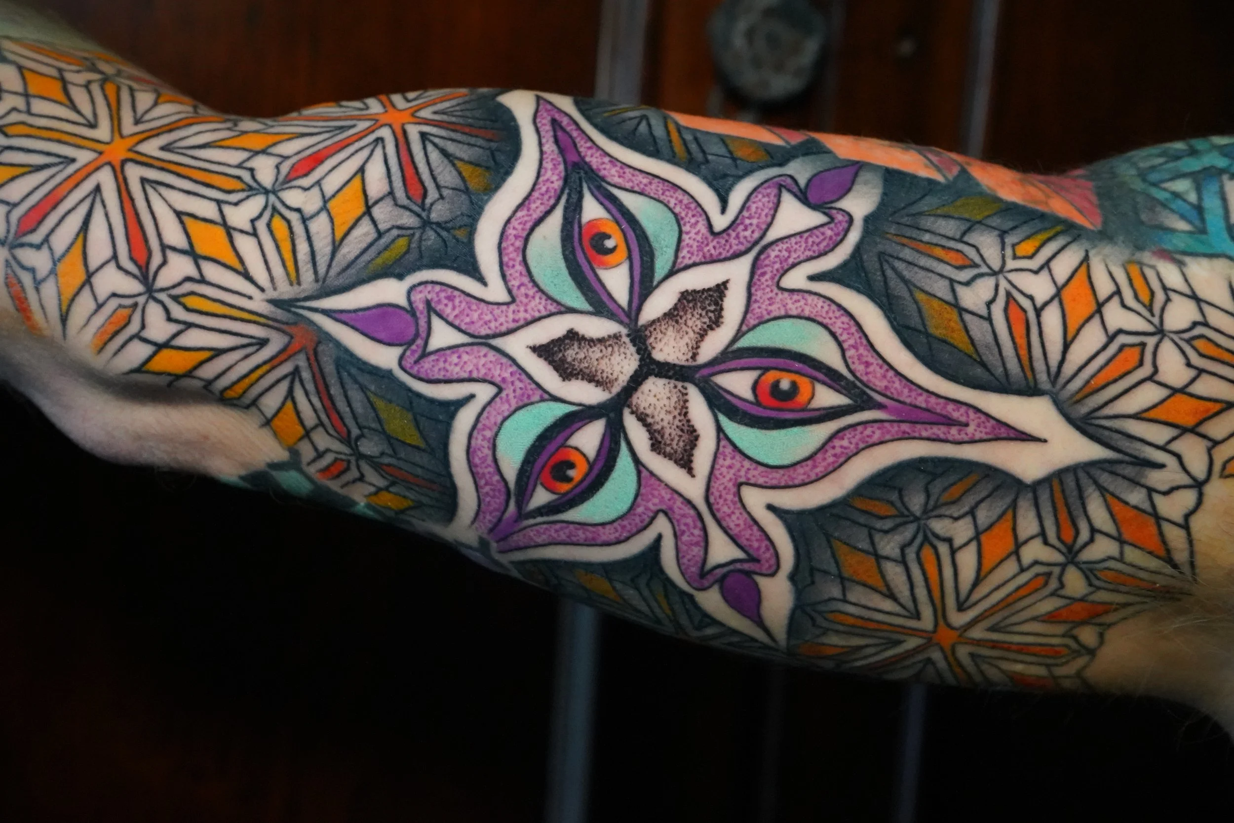 Colorful geometric tattoo design featuring a central butterfly with four eyes, surrounded by intricate star and hexagon patterns.
