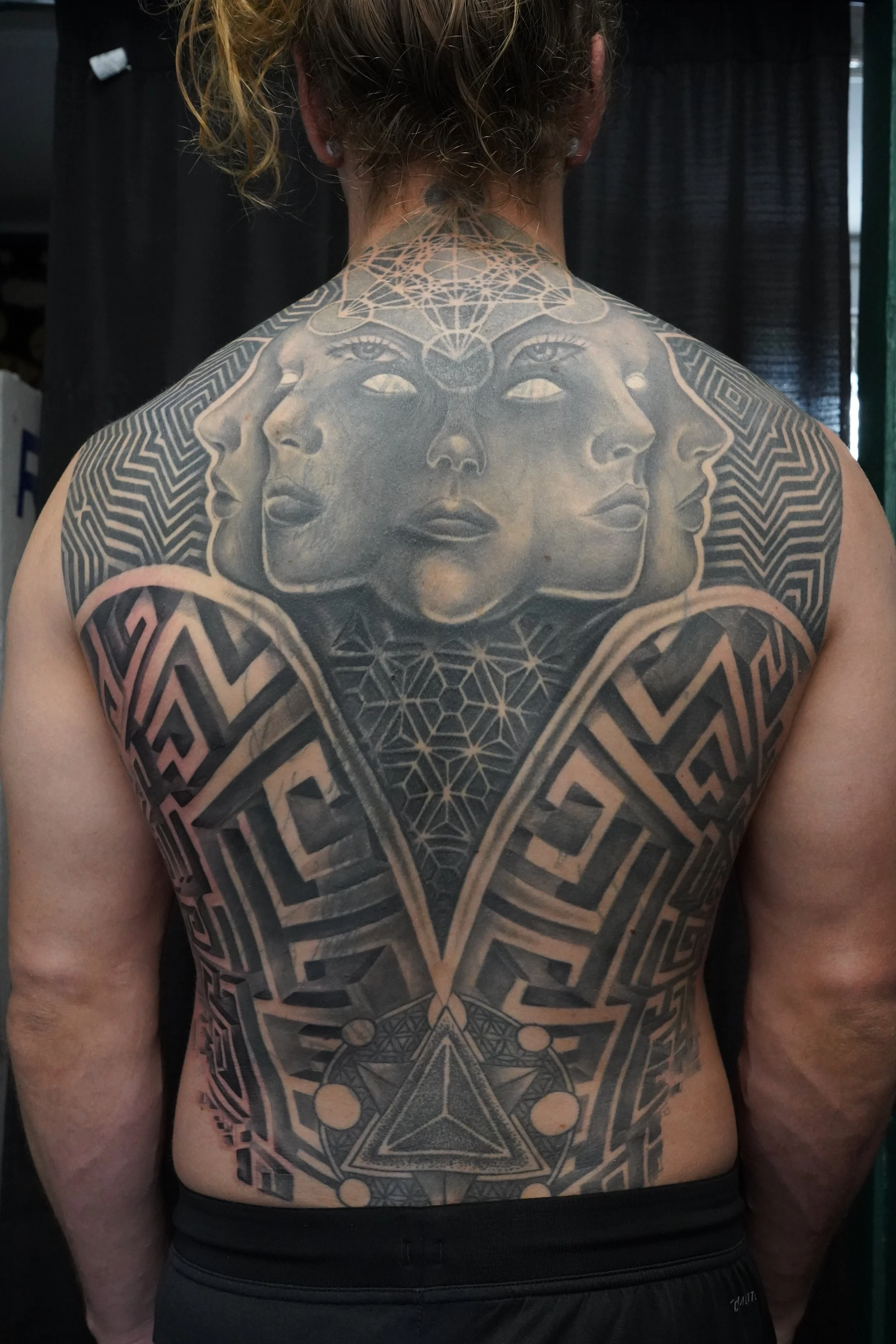 A person's back with a large black geometric and surreal tattoo featuring multiple faces, eyes, and celestial symbols.