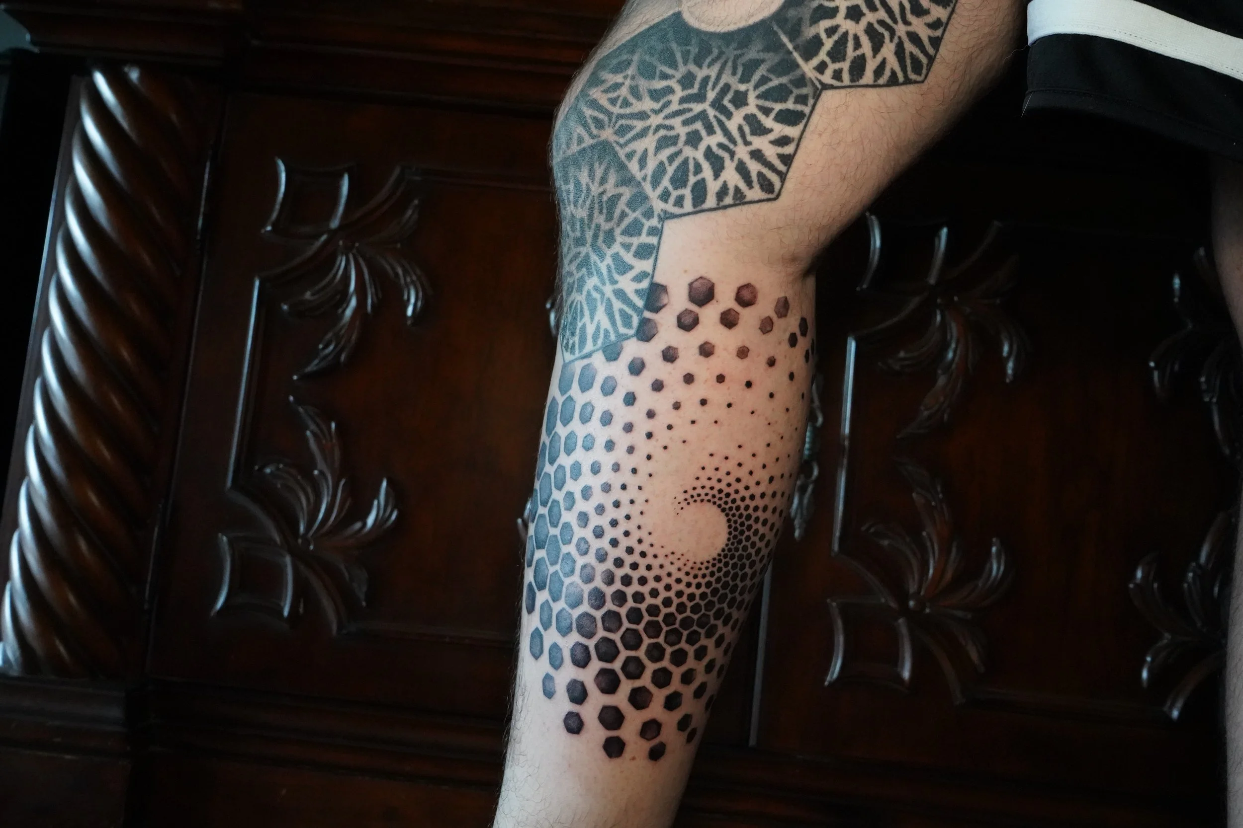 Tattooed arm with geometric and dotted circle patterns, resting on a wooden carved surface.