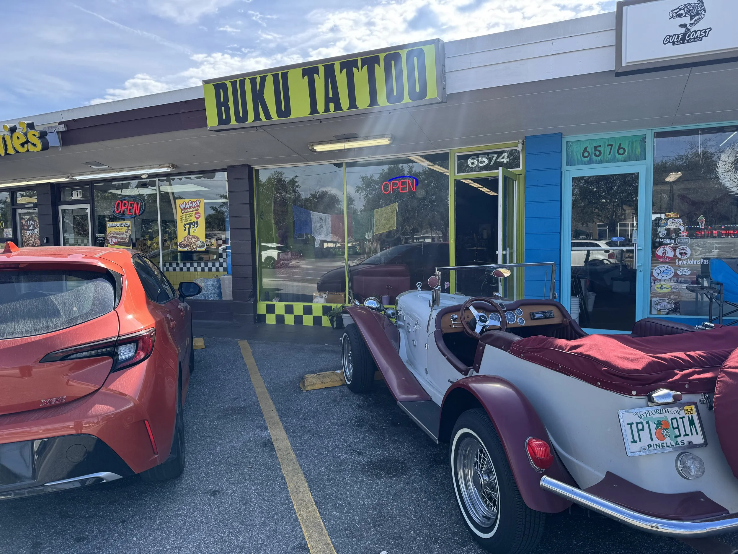 A parking lot in front of a tattoo shop named 'Buku Tattoo' with a red modern car and a vintage white and maroon tricycle-style car parked outside. The tattoo shop has a green and black sign, and neon 'Open' signs in the windows.