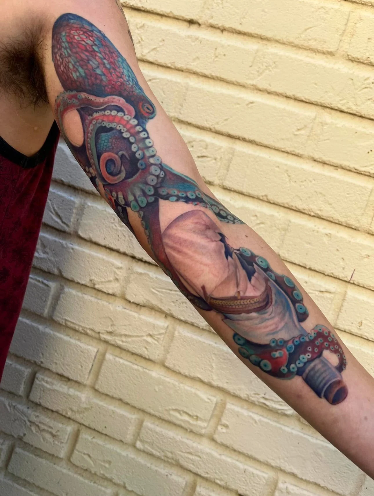 Colorful tattoo of an octopus and a ship on a person's arm against a brick wall.