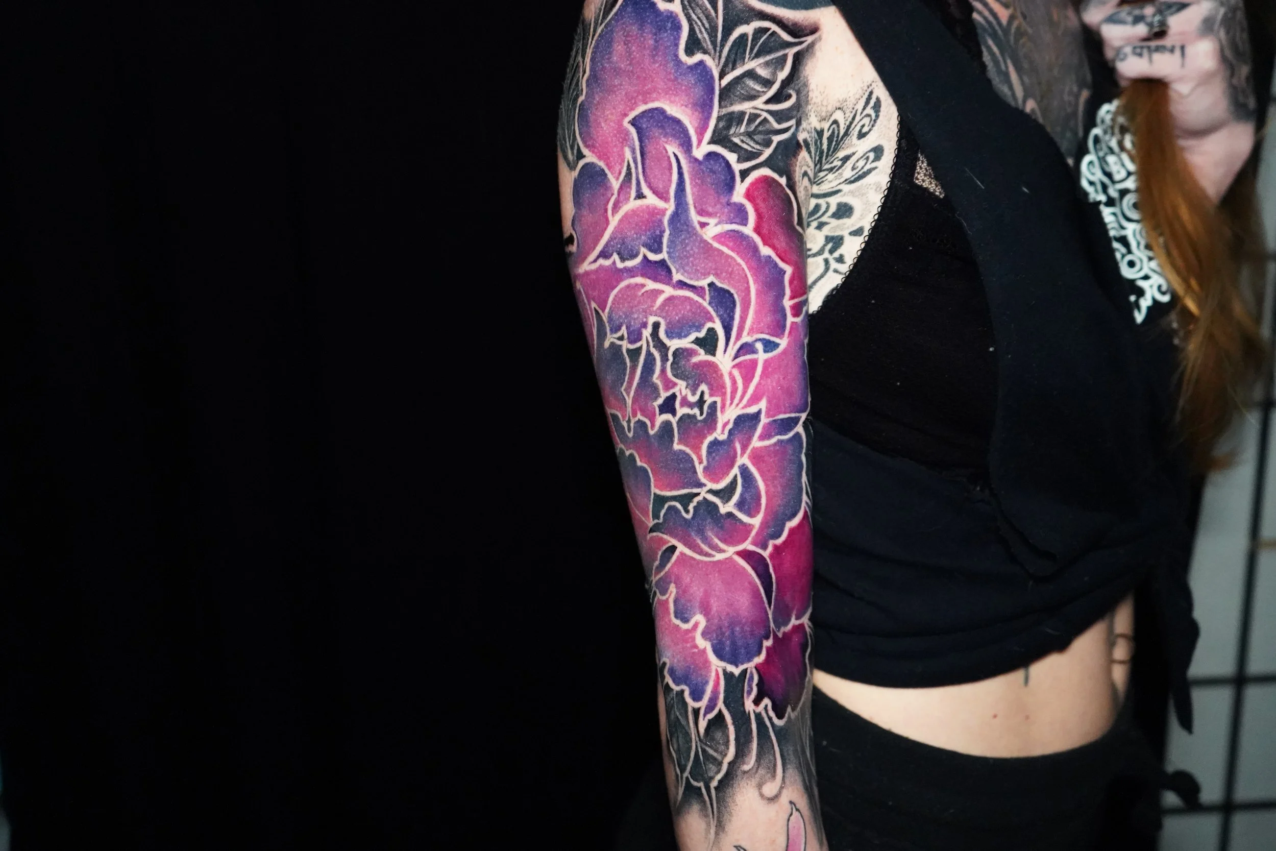 Colorful floral tattoo on woman's upper arm, with purple and pink flowers outlined in white, against a black background.