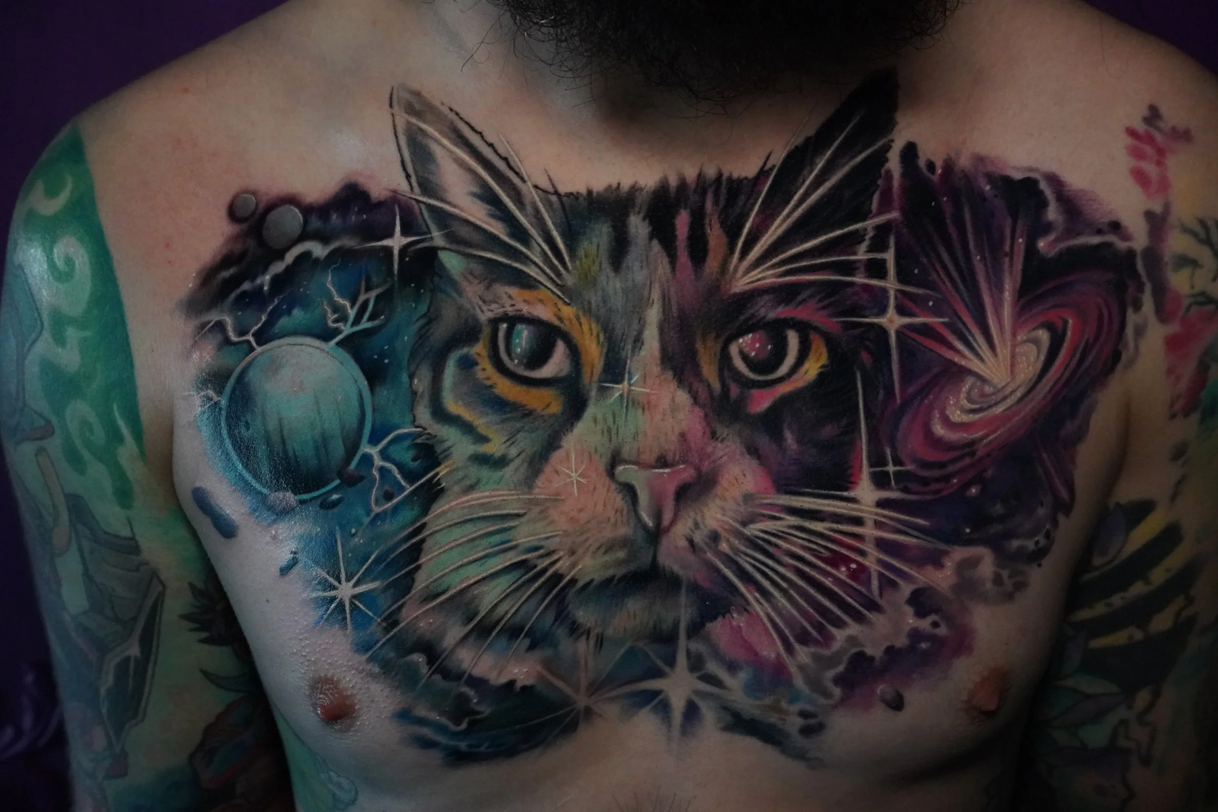 Colorful tattoo of a cat's face with a cosmic and galaxy theme, including planets, stars, and swirling patterns, on a person's chest and shoulders.
