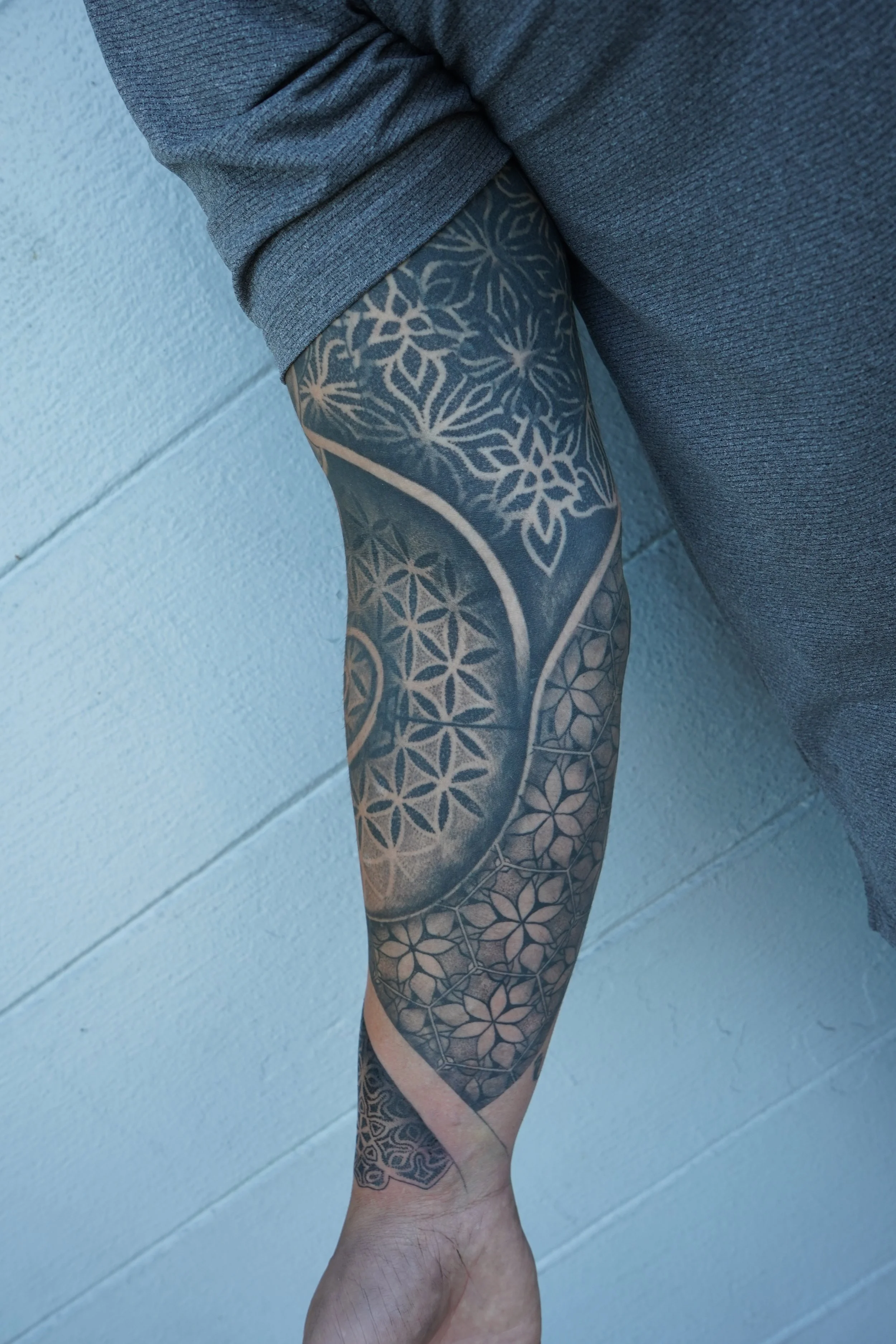 Close-up of a person's arm with intricate black and gray geometric tattoos, extending from the shoulder to the wrist, with a person wearing a gray sleeve rolled up to reveal the tattoos, against a pale blue wall.