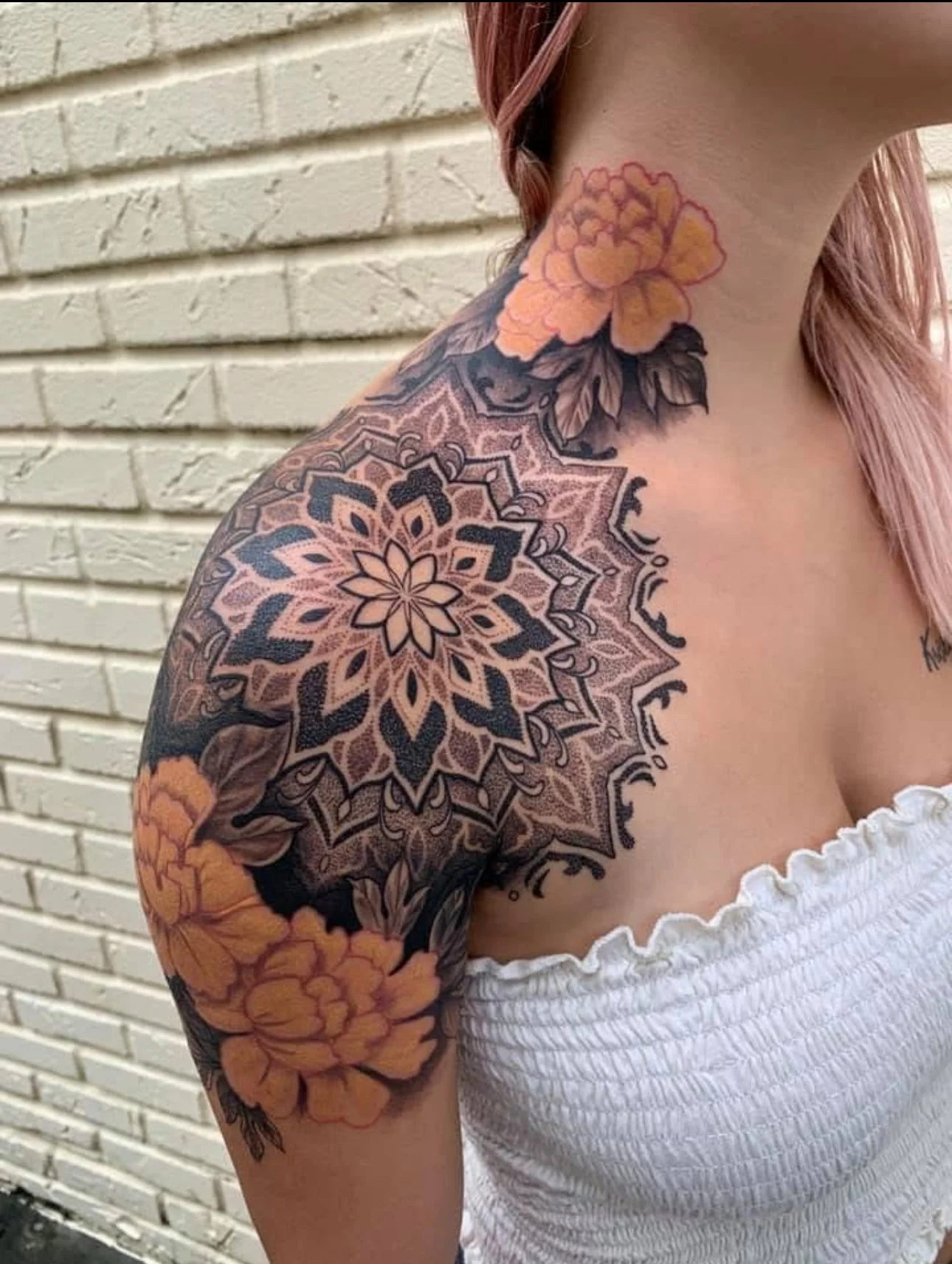 A woman with pink hair has a detailed floral tattoo covering her shoulder, upper arm, and neck. The tattoo includes large orange flowers, dark leaves, and an intricate mandala pattern with pink and black details on her shoulder.