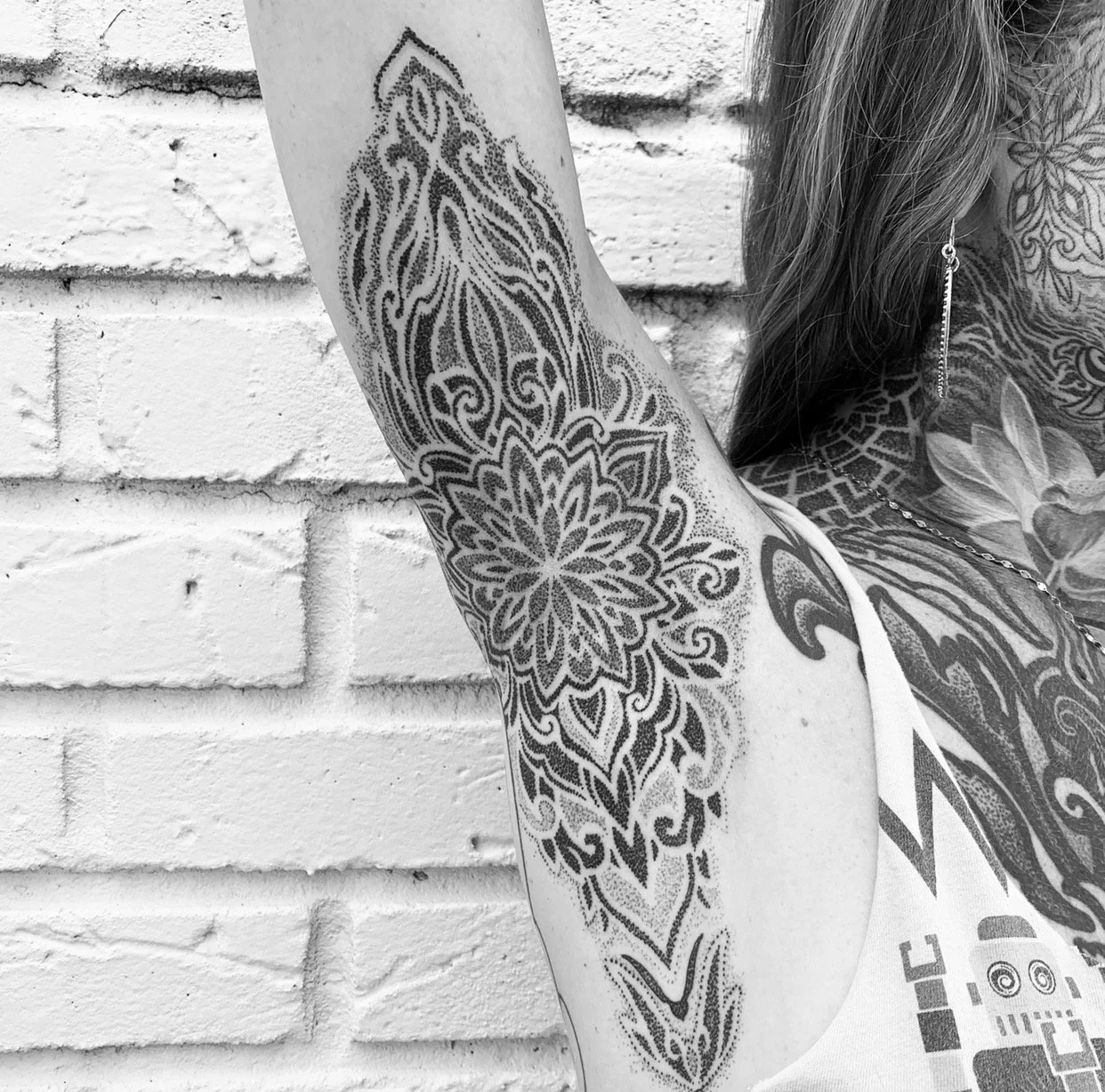 A person with extensive tattoos on their arm, shoulder, and neck, standing against a white brick wall, wearing an earring and a tank top.