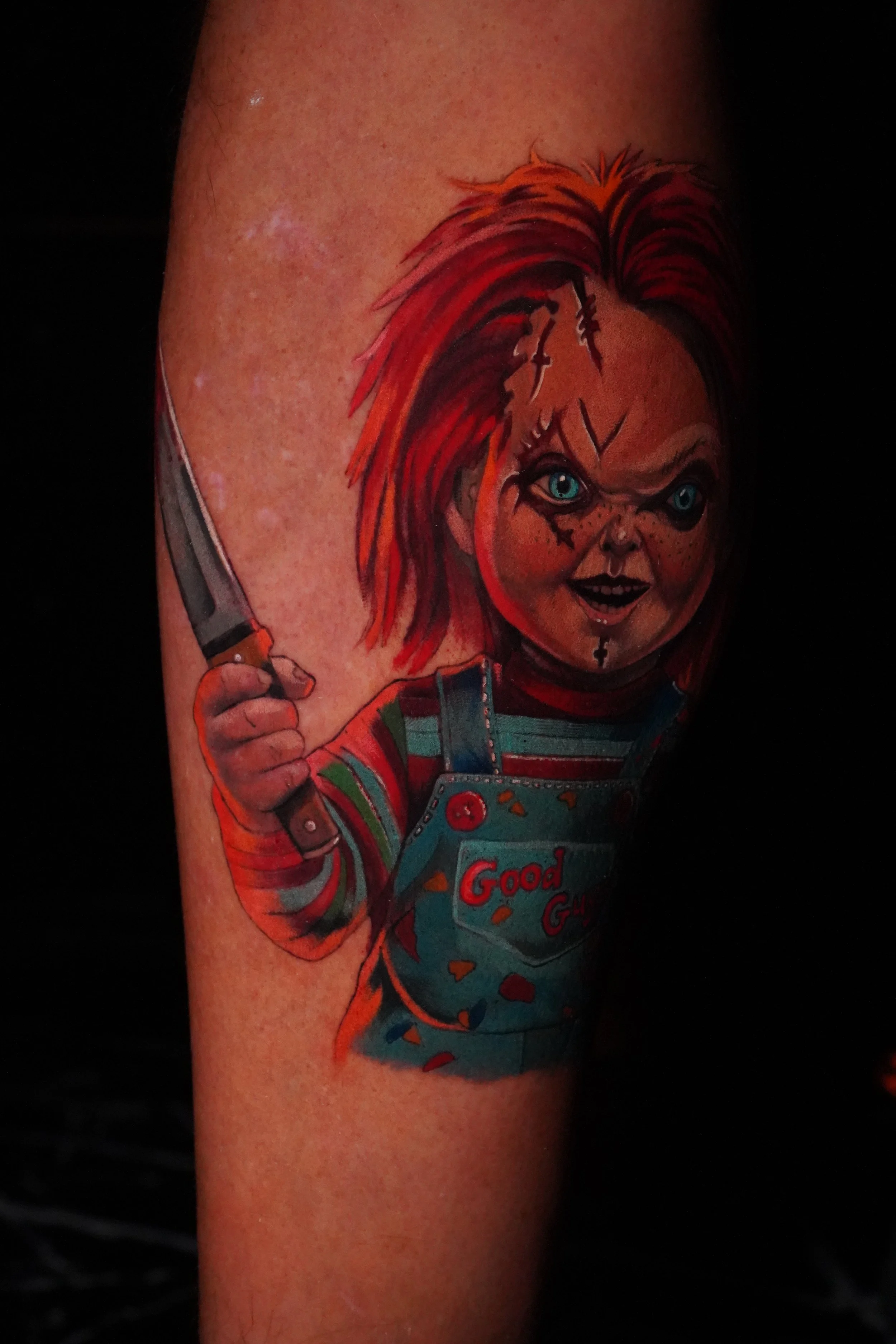 Tattoo of Chucky, the doll from the horror movie, holding a knife and wearing overalls with the words 'Good G**' on it.