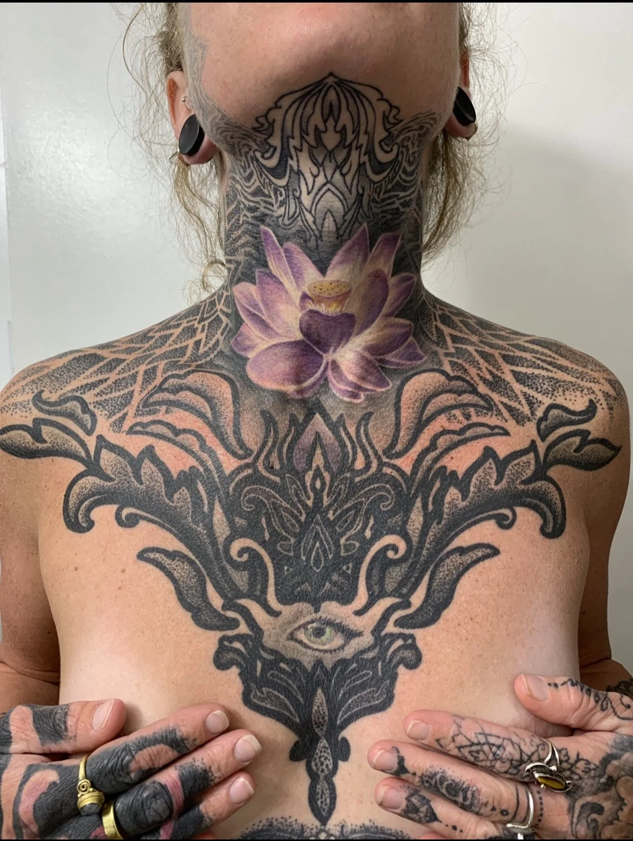 A person with extensive tattoos, including a large lotus flower tattoo on the neck, a detailed black tribal tattoo on the chest, and a tattooed eye on the stomach. They have stretched earlobes with black tunnel gauges and multiple rings and tattoos o