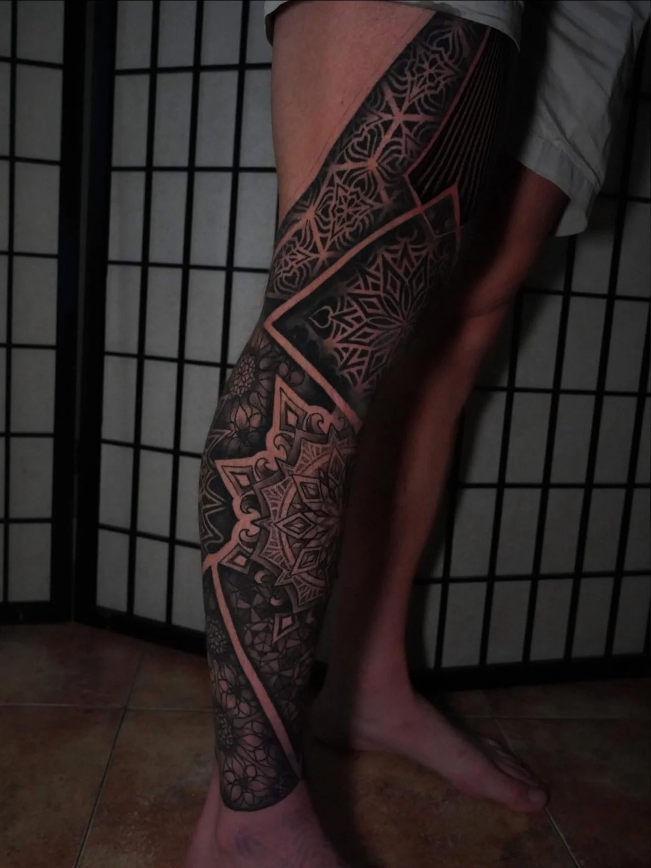 A person's leg with an intricate black and pink geometric tattoo design covering from thigh to ankle, standing against a black grid background.