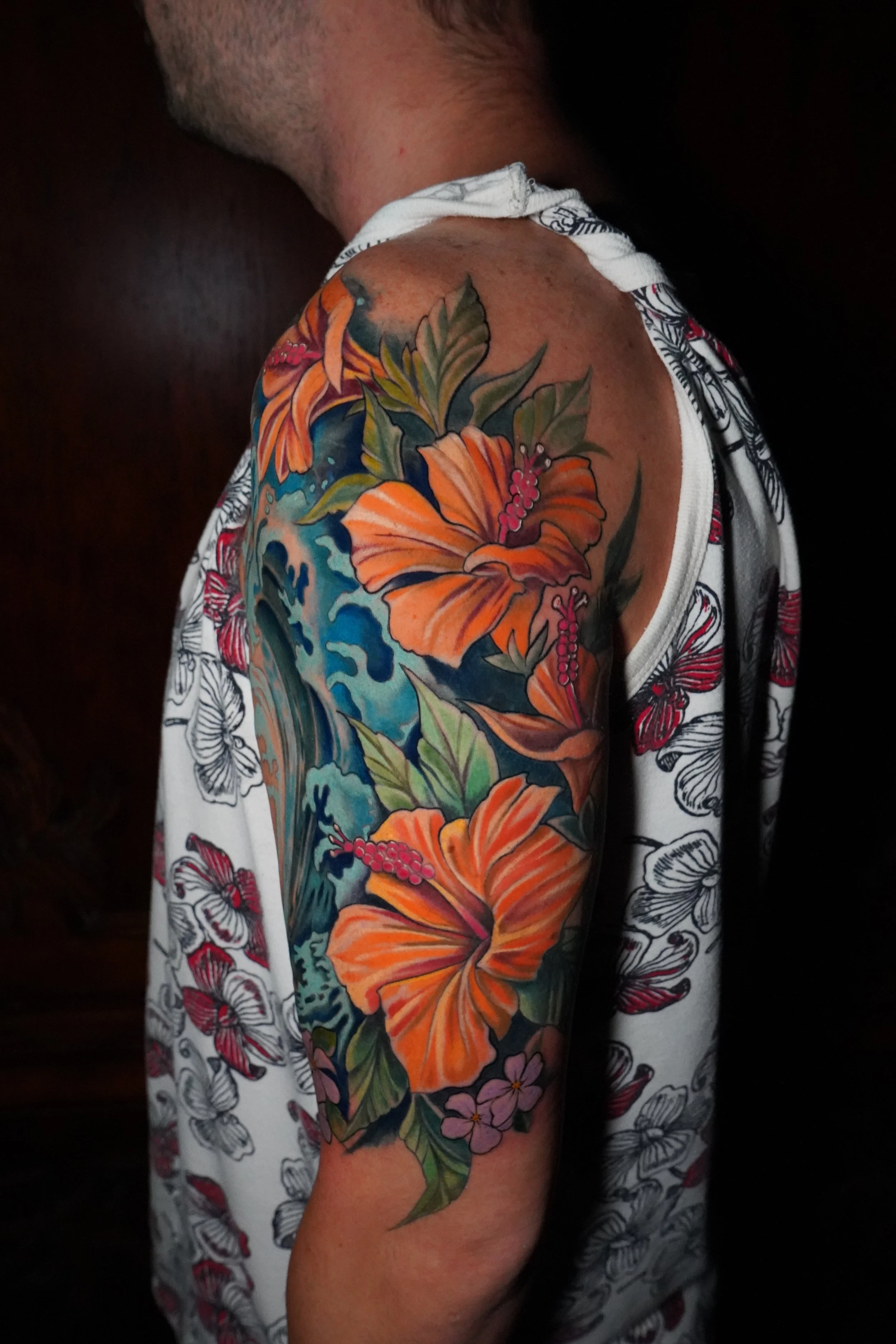 A person's upper arm and shoulder with a colorful hibiscus flower tattoo and green leaves, featuring shades of orange, pink, and green, against a dark background.