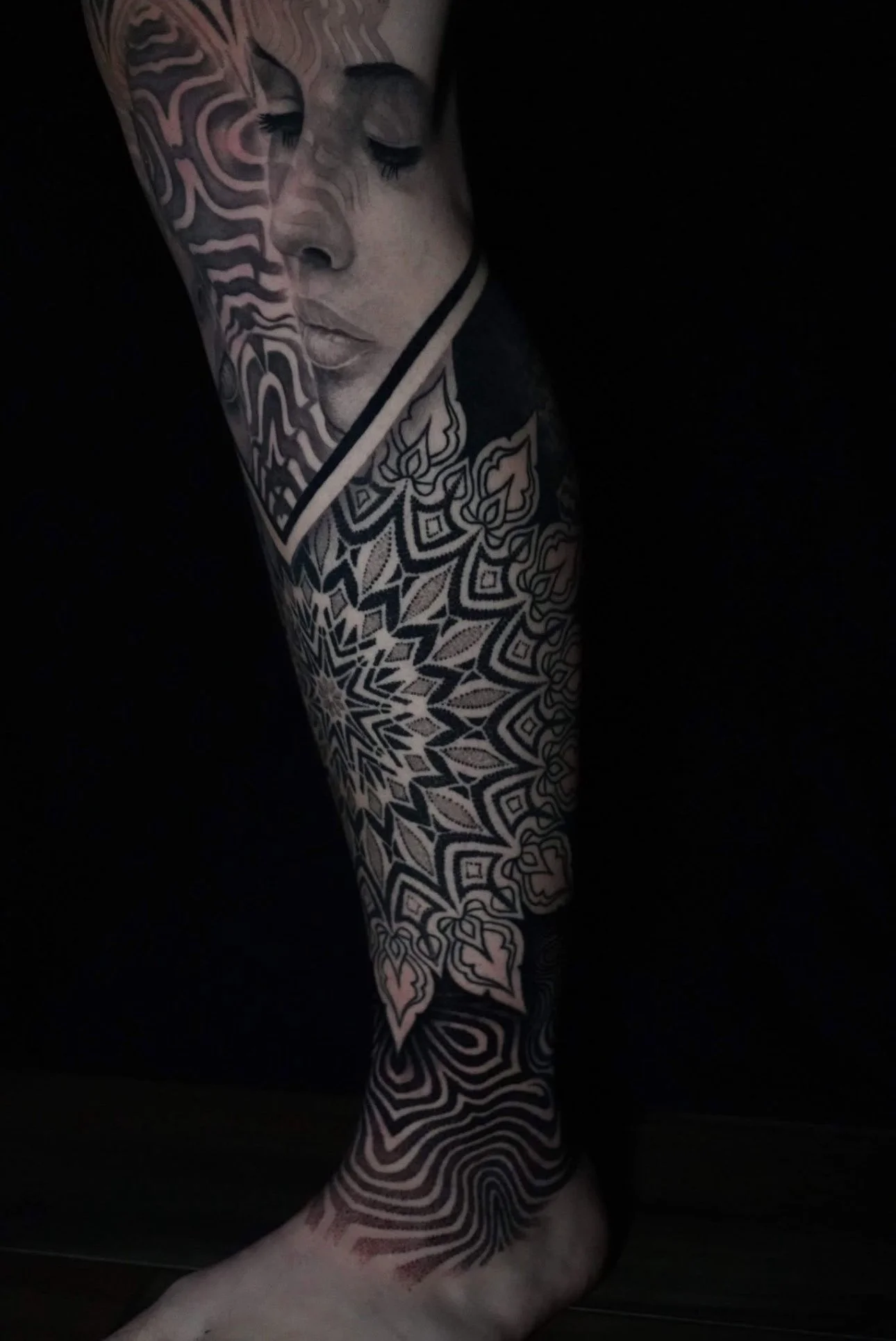 Close-up of a person's leg with intricate black and gray tattoos, including a portrait of a woman's face with closed eyes and detailed patterns covering the leg.