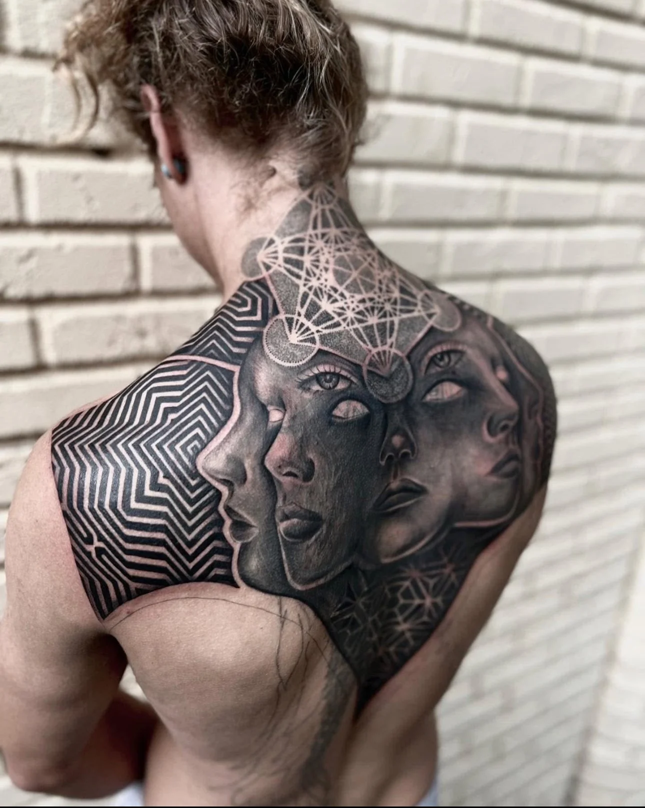 A person with short curly hair and tattoos on their back and shoulder, standing against a brick wall. The tattoos feature artistic faces, geometric patterns, and a network of interconnected lines and shapes.