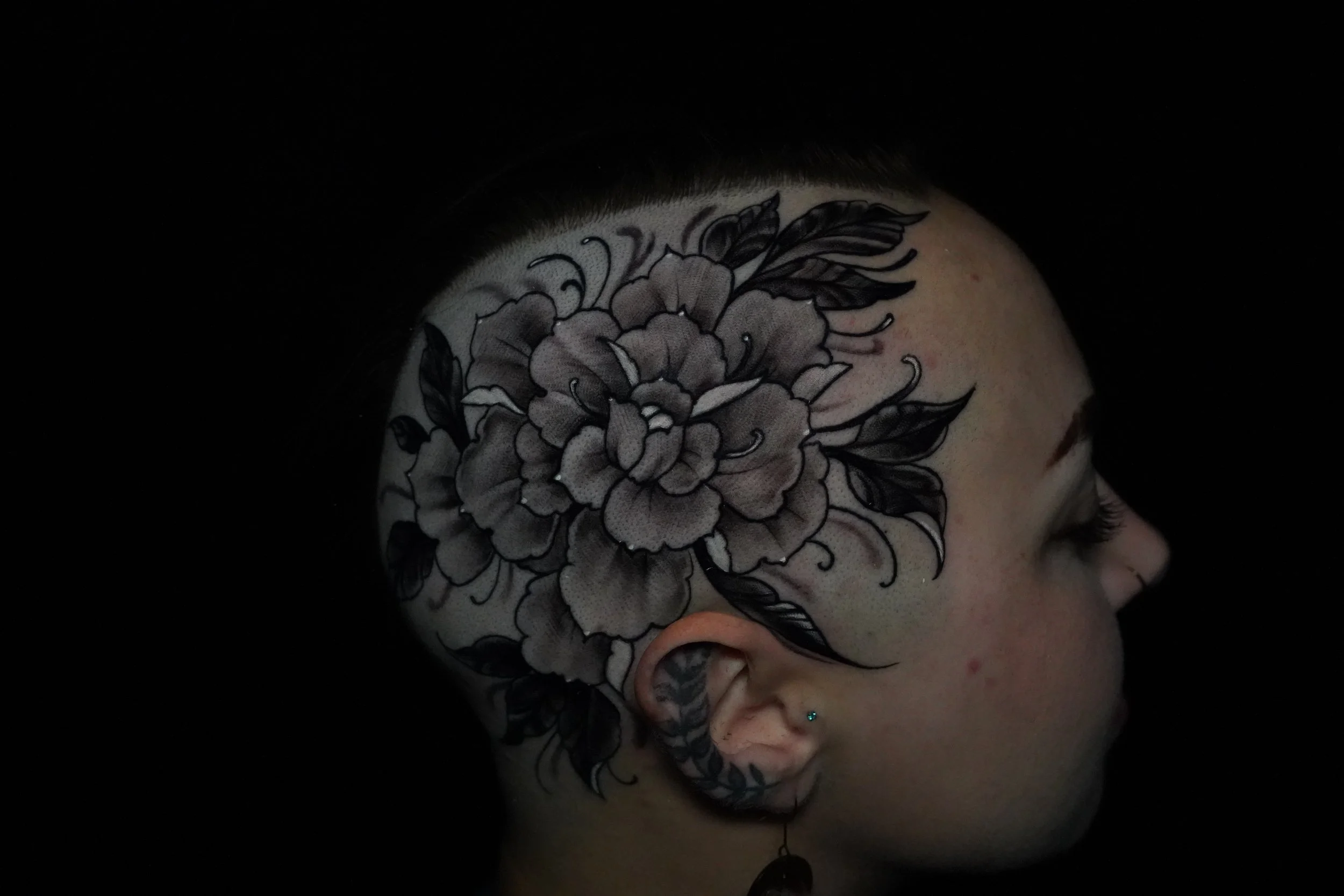 A person with a detailed black and gray floral tattoo on the side of their head, featuring a large flower and leaves, against a dark background.