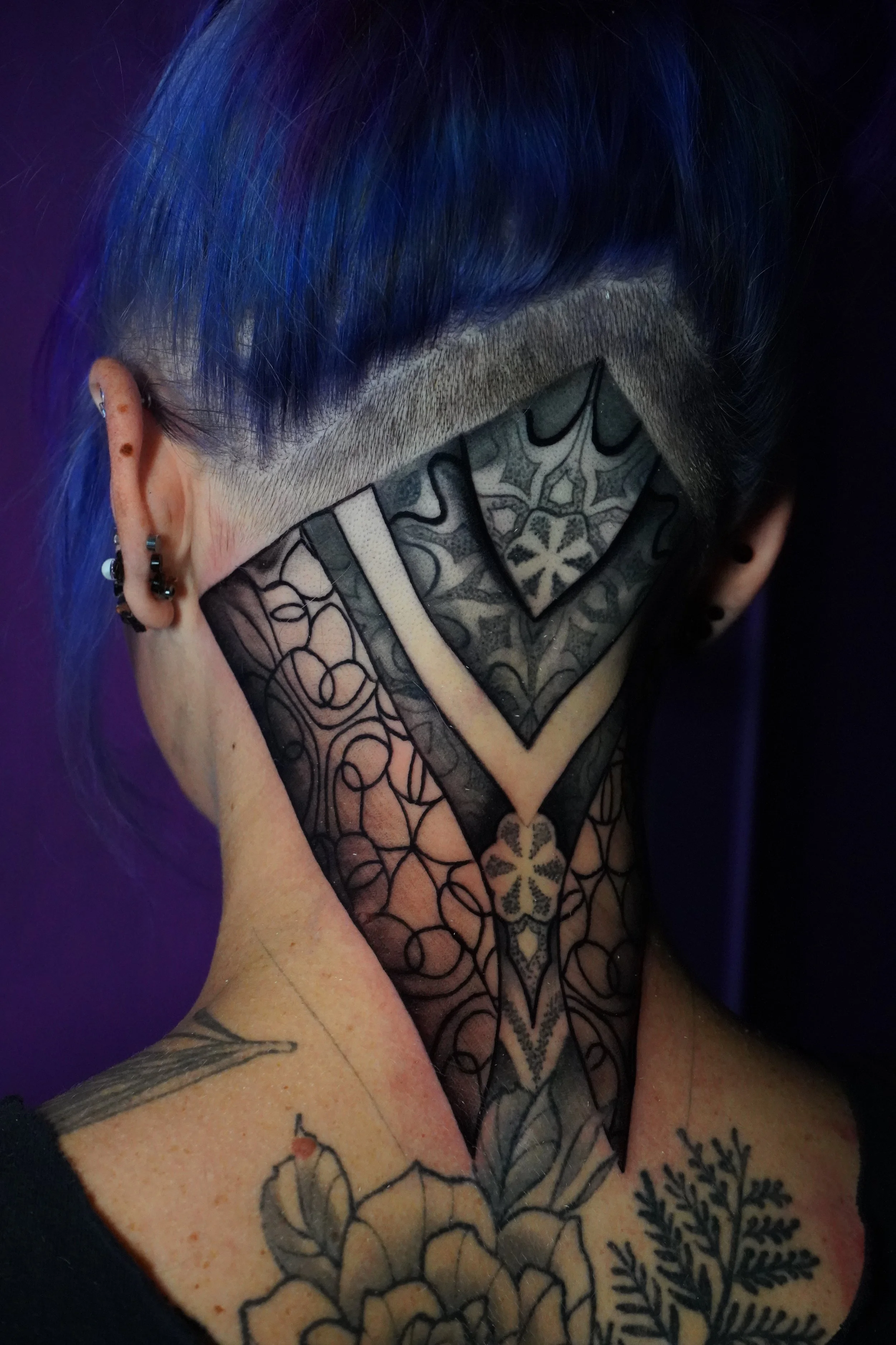 Person with blue hair and multiple earrings showing a large black and gray geometric tattoo covering the neck and part of the face, along with tattoos on the shoulder.