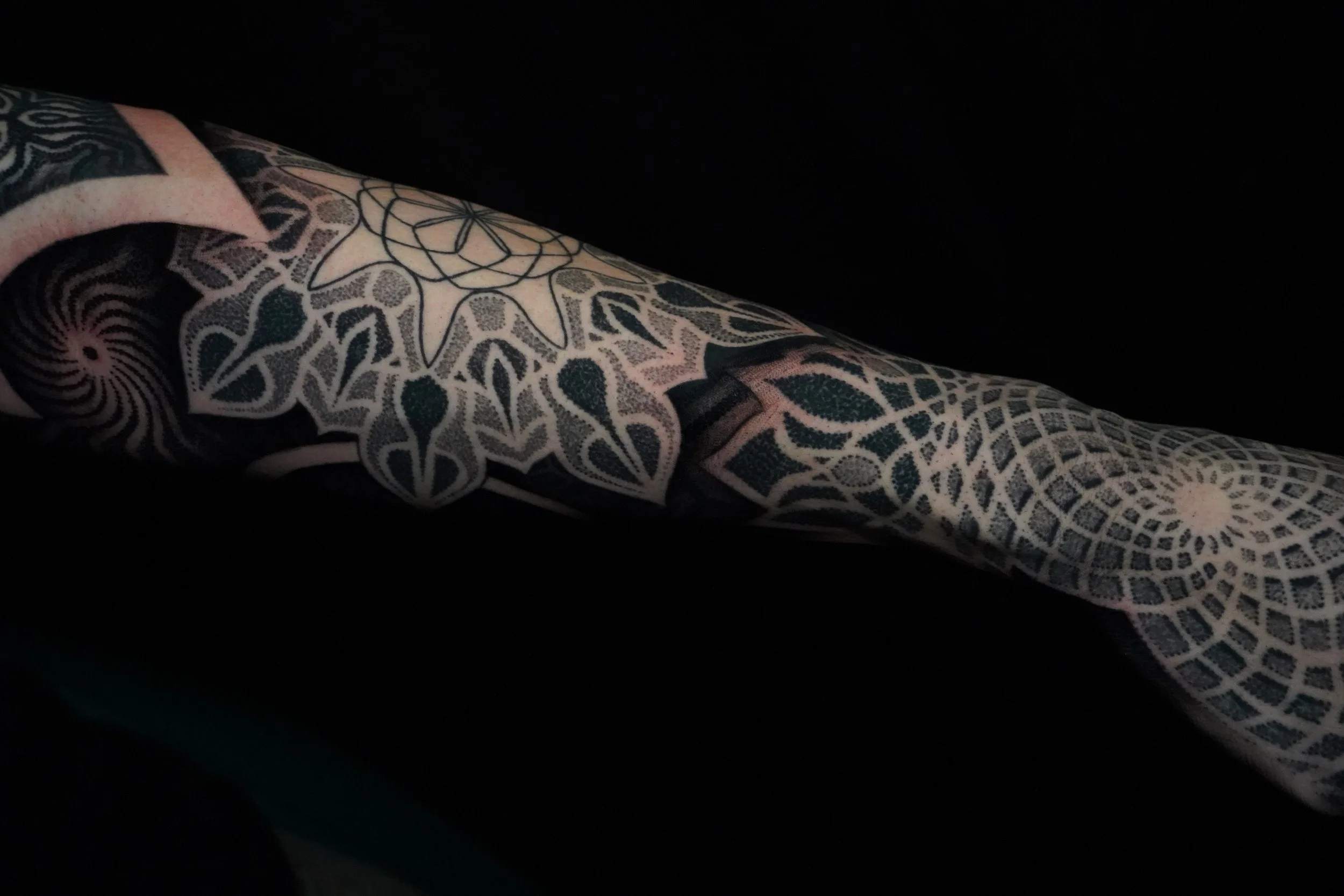 Close-up of a tattooed arm with intricate geometric and floral black ink designs against a black background.
