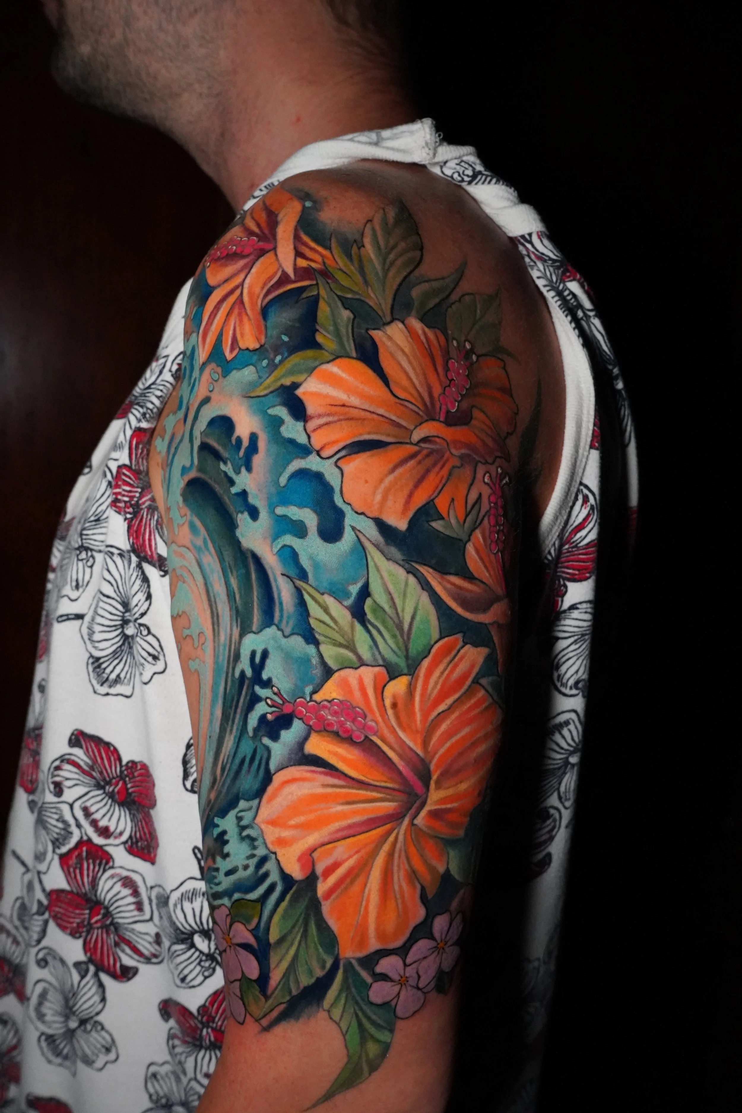 Colorful tattoo of orange hibiscus flowers, green leaves, pink berries, and a blue wave on a man's upper arm.