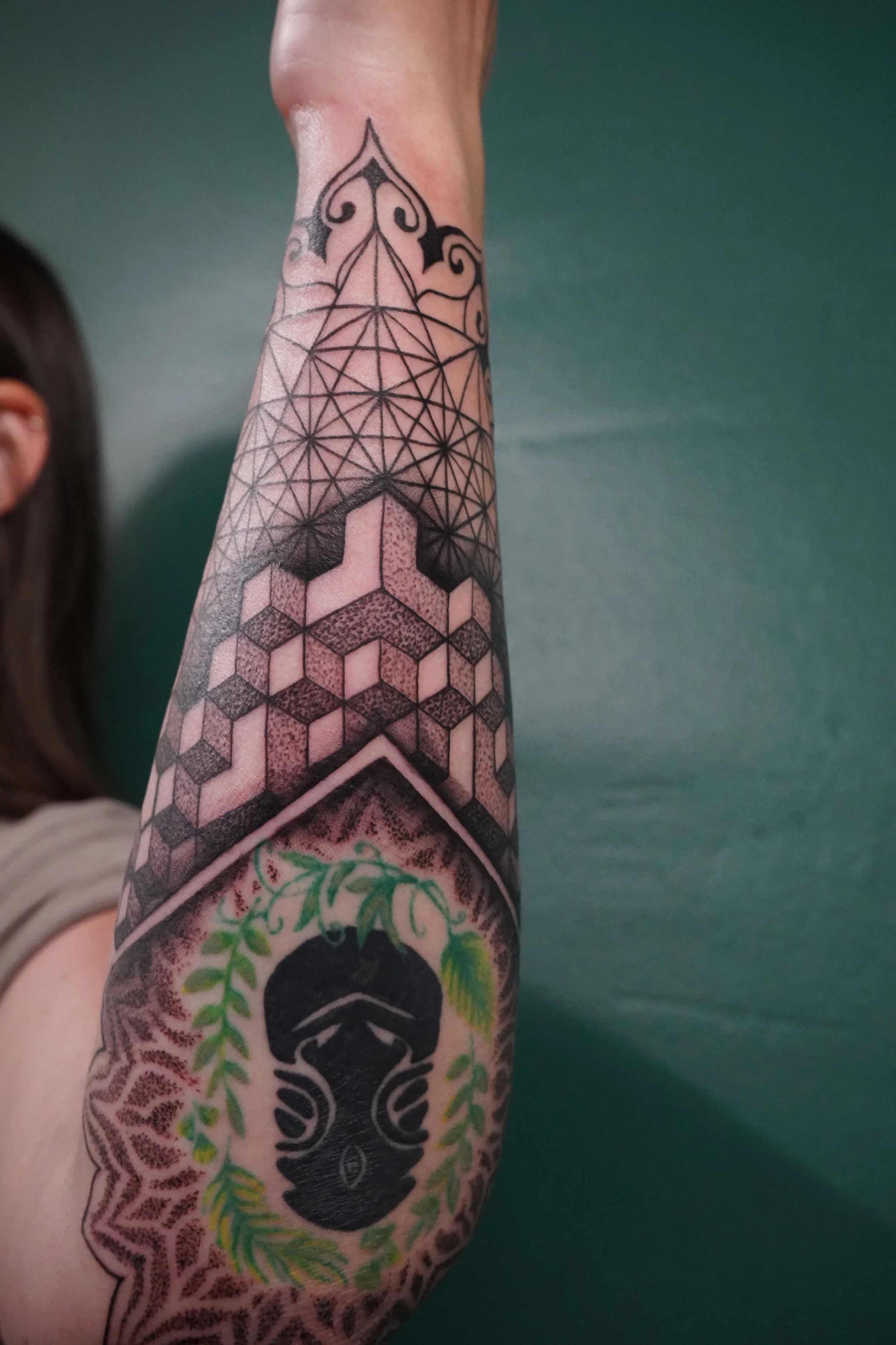 Tattoo of a black gas mask with green leaves and vines on the forearm, with geometric and abstract patterns extending up the arm.