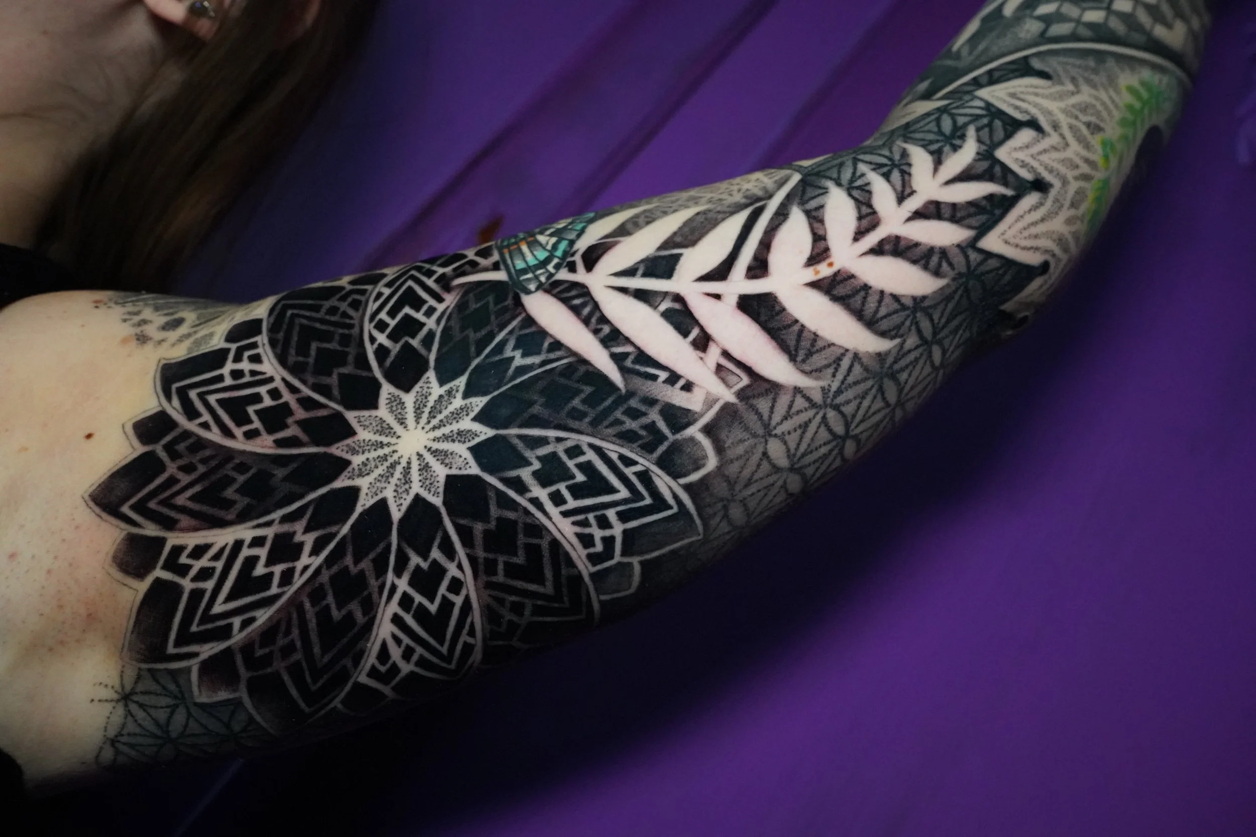 Close-up of a person's arm with a black and white geometric and floral tattoo, lying on a purple surface.
