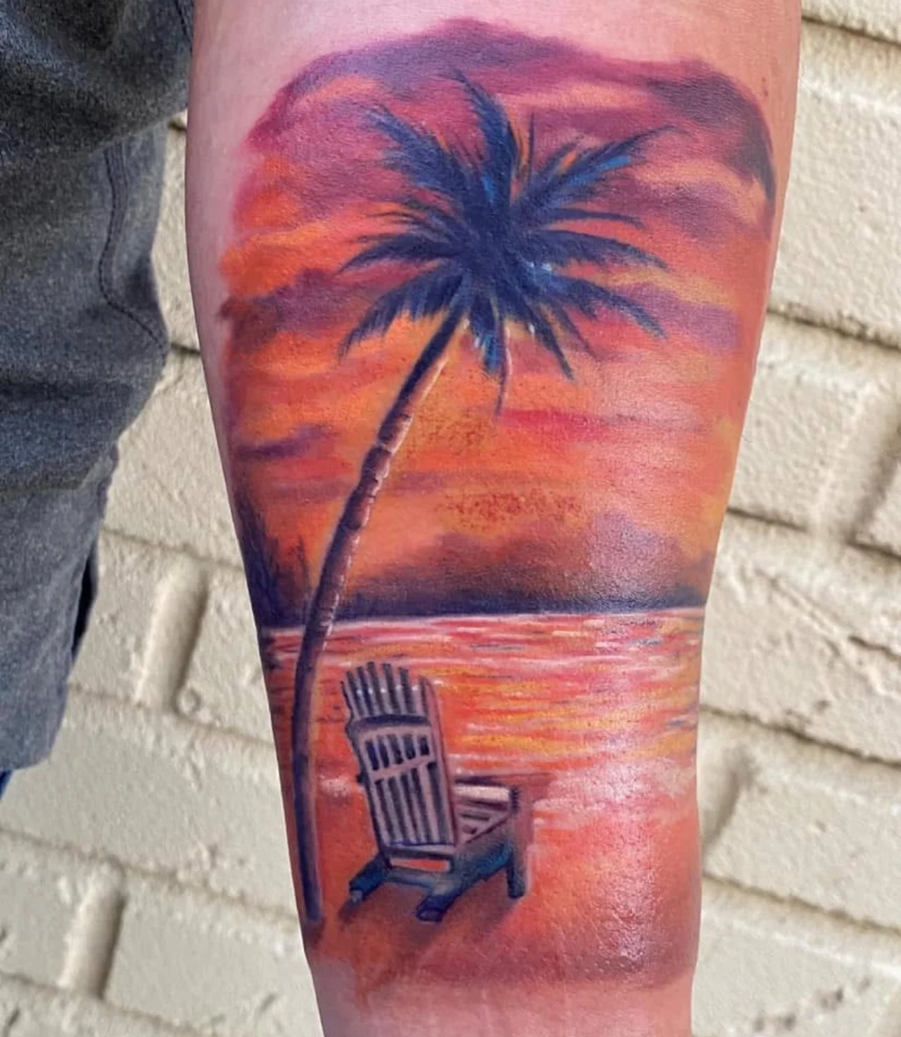 Colorful tattoo of a sunset scene with a palm tree and an empty beach chair on the arm.