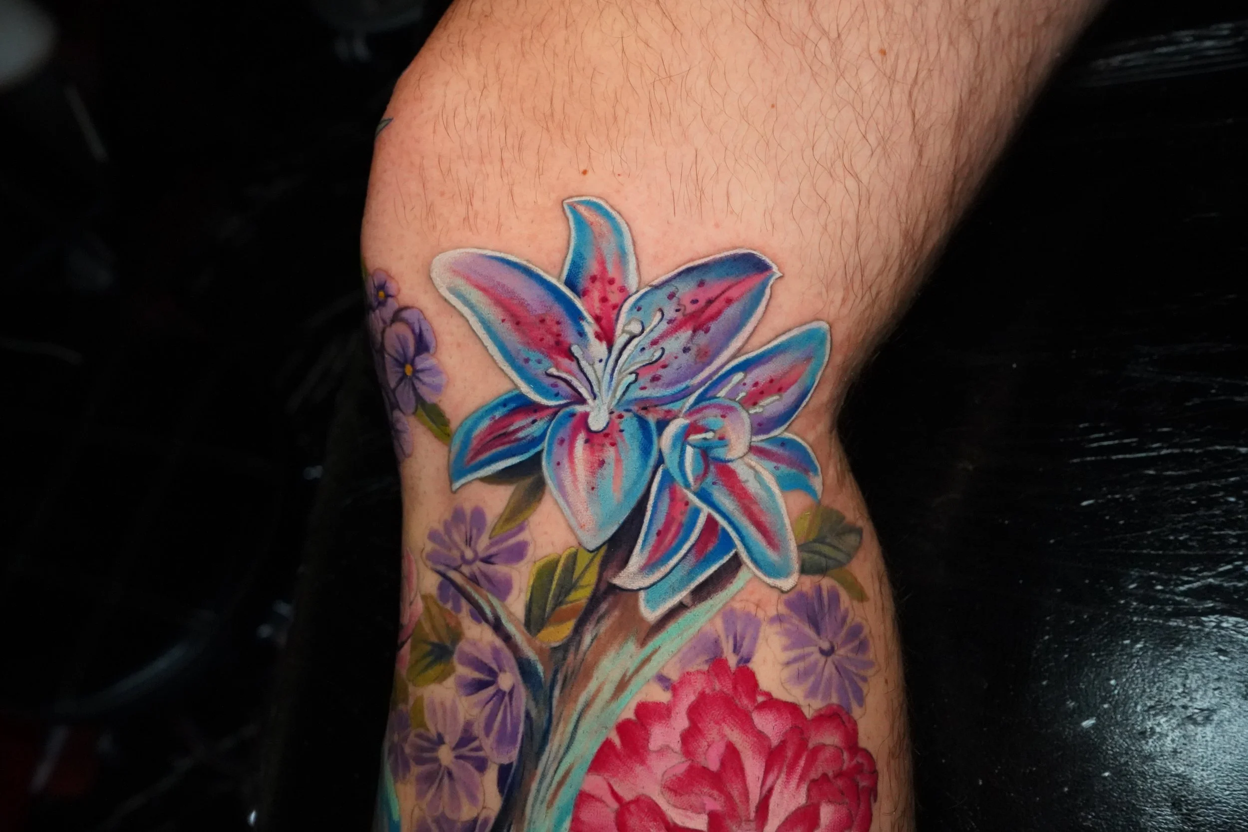 Colorful tattoo of a large blue and pink lily flower on a person's arm, surrounded by smaller purple flowers and green leaves.