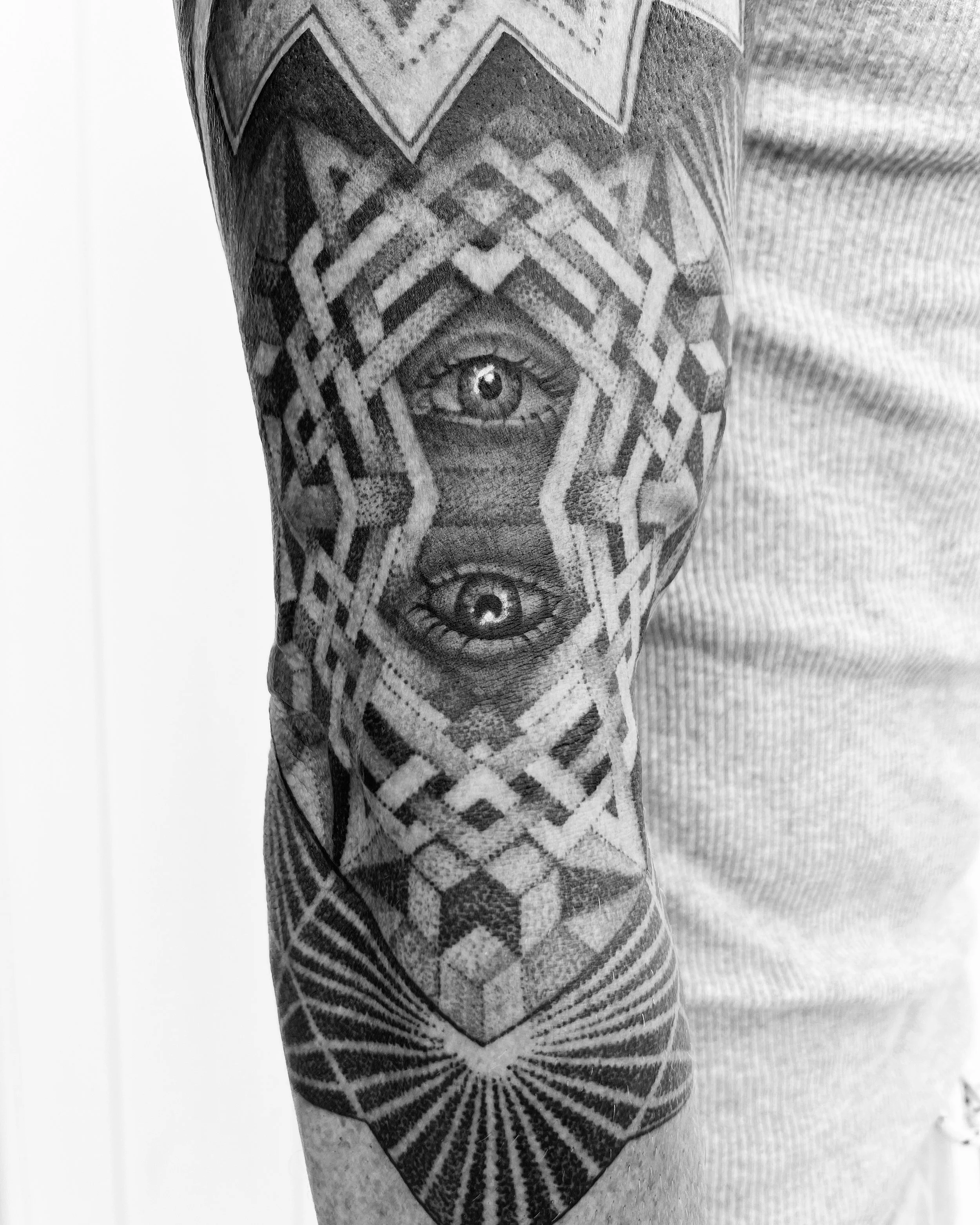 Black and white photo of a tattooed arm with geometric patterns and two detailed human eyes tattooed on the inner side.