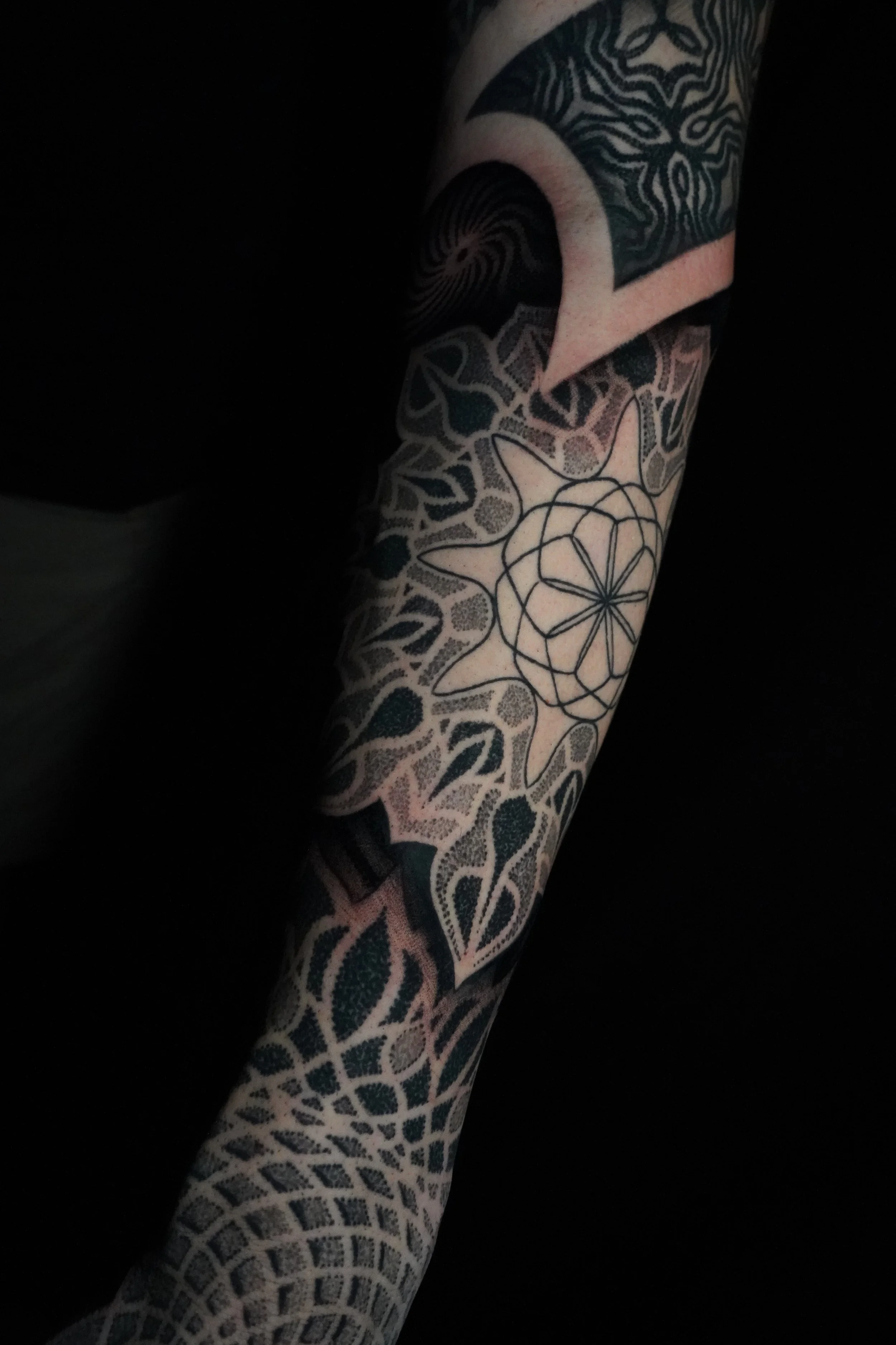 A tattooed arm with geometric and abstract designs, including a central star-shaped pattern surrounded by a web-like circle, leaves, and wavy lines, on a dark background.