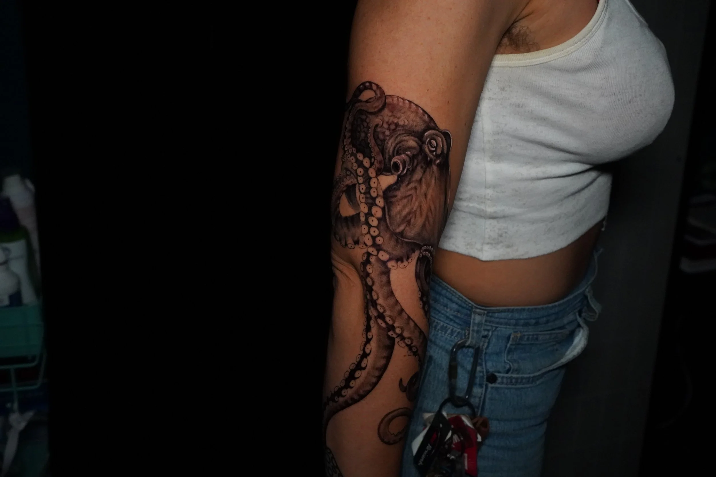 A person's upper arm with a detailed octopus and lion face tattoo, wearing a white sleeveless shirt and blue jeans.