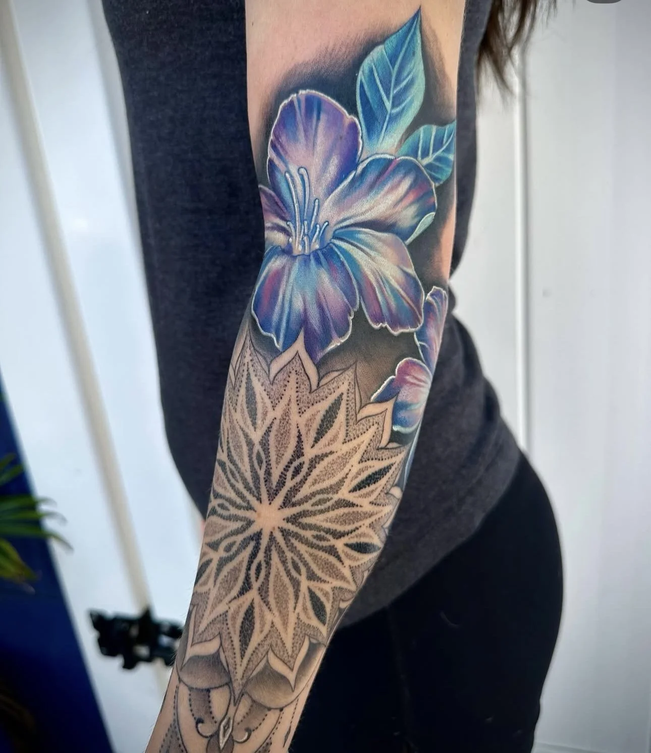 Close-up of a colorful tattoo on a person's arm featuring a large purple-blue flower with green leaves and a geometric pattern below.
