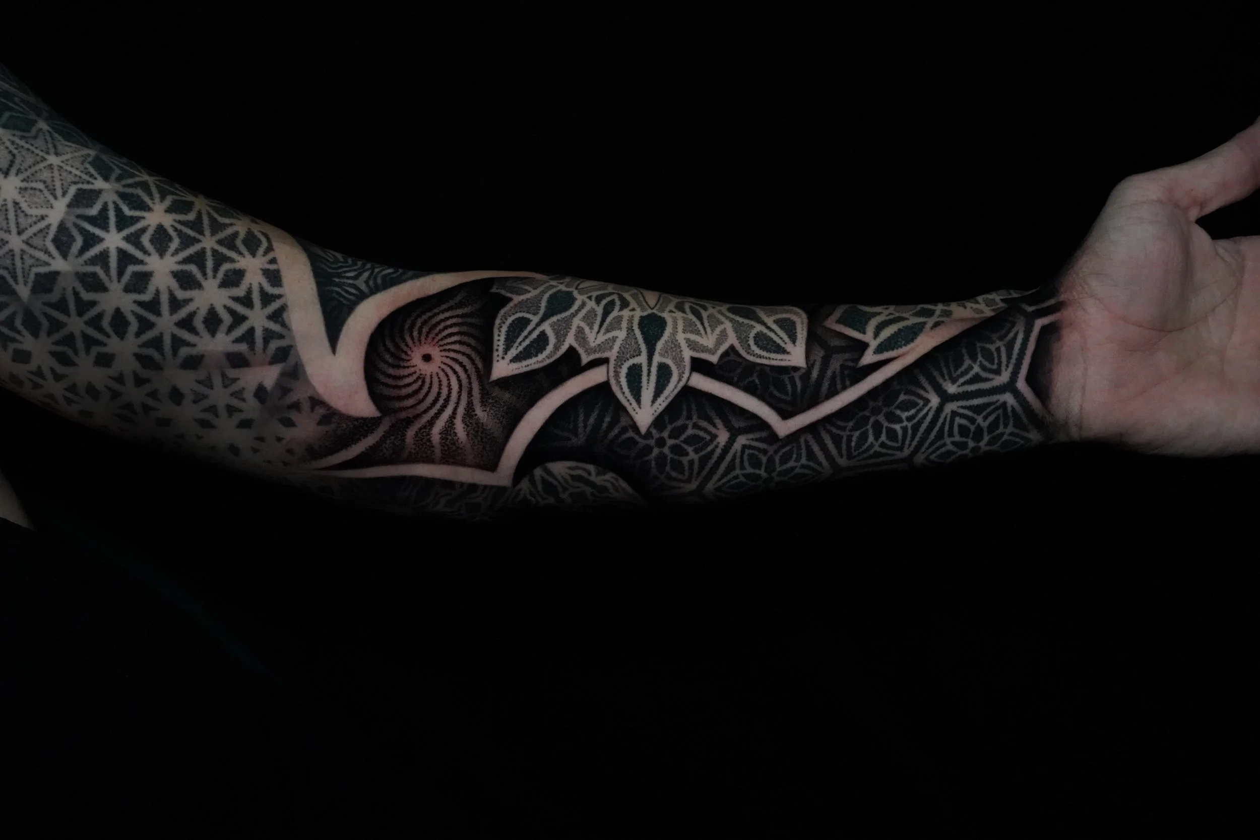 Close-up of a person's arm with a detailed geometric black and gray tattoo sleeve that includes a spiral pattern, floral motifs, and interconnected shapes, against a black background.