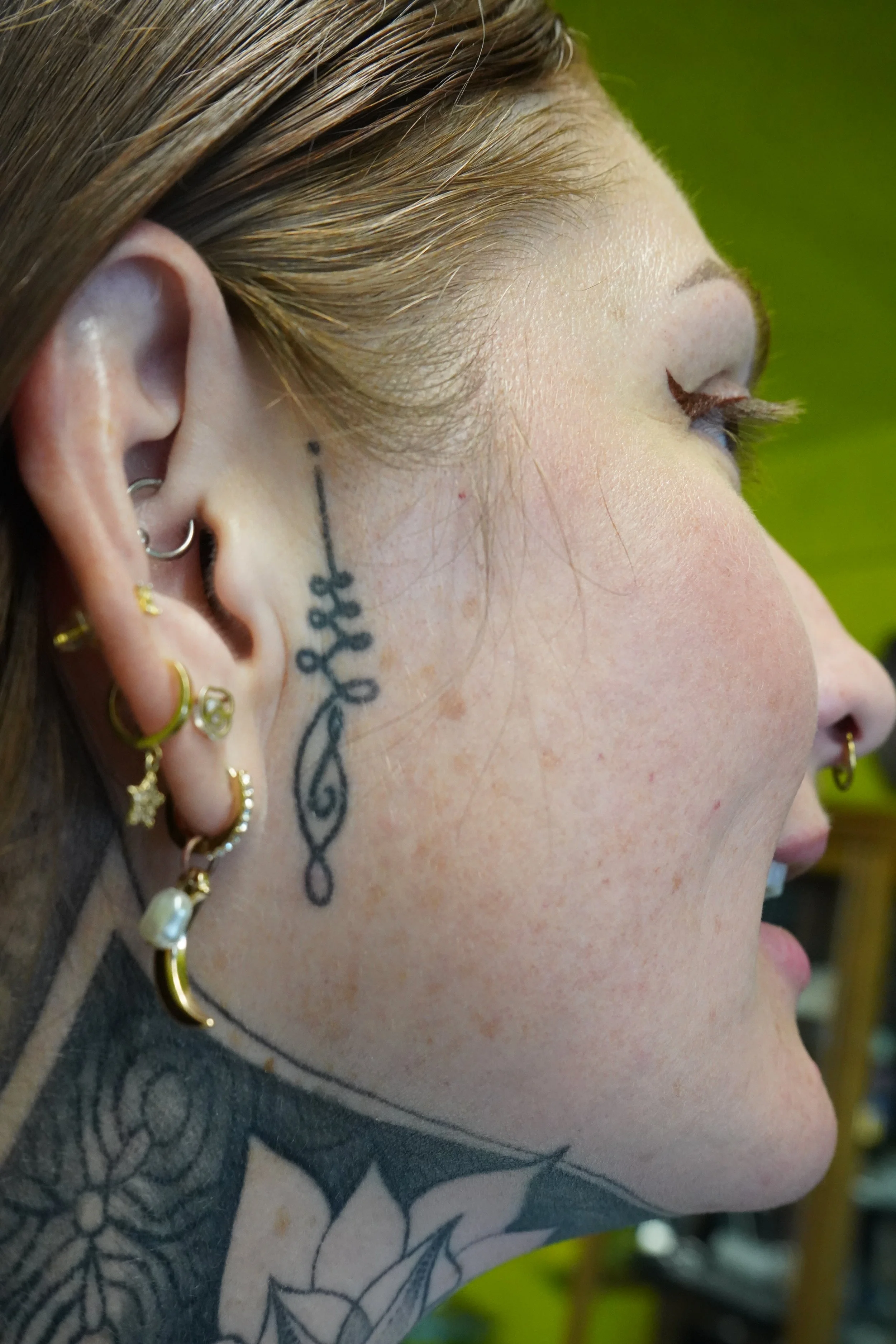 Close-up of a person's face with multiple earrings and a tattoo behind the ear, showing fair skin with freckles, hair pulled back, and a green background.