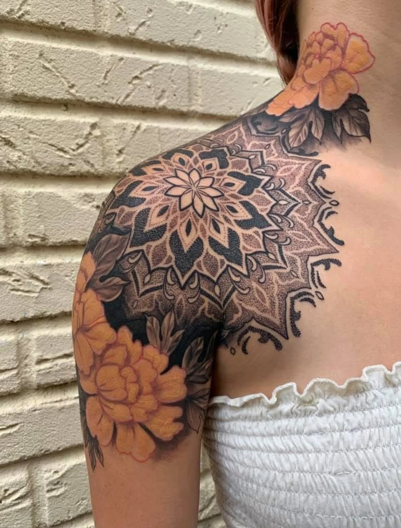 Close-up of a shoulder tattoo featuring a large, intricate mandala design with floral elements, complemented by orange flowers on the upper arm and shoulder.
