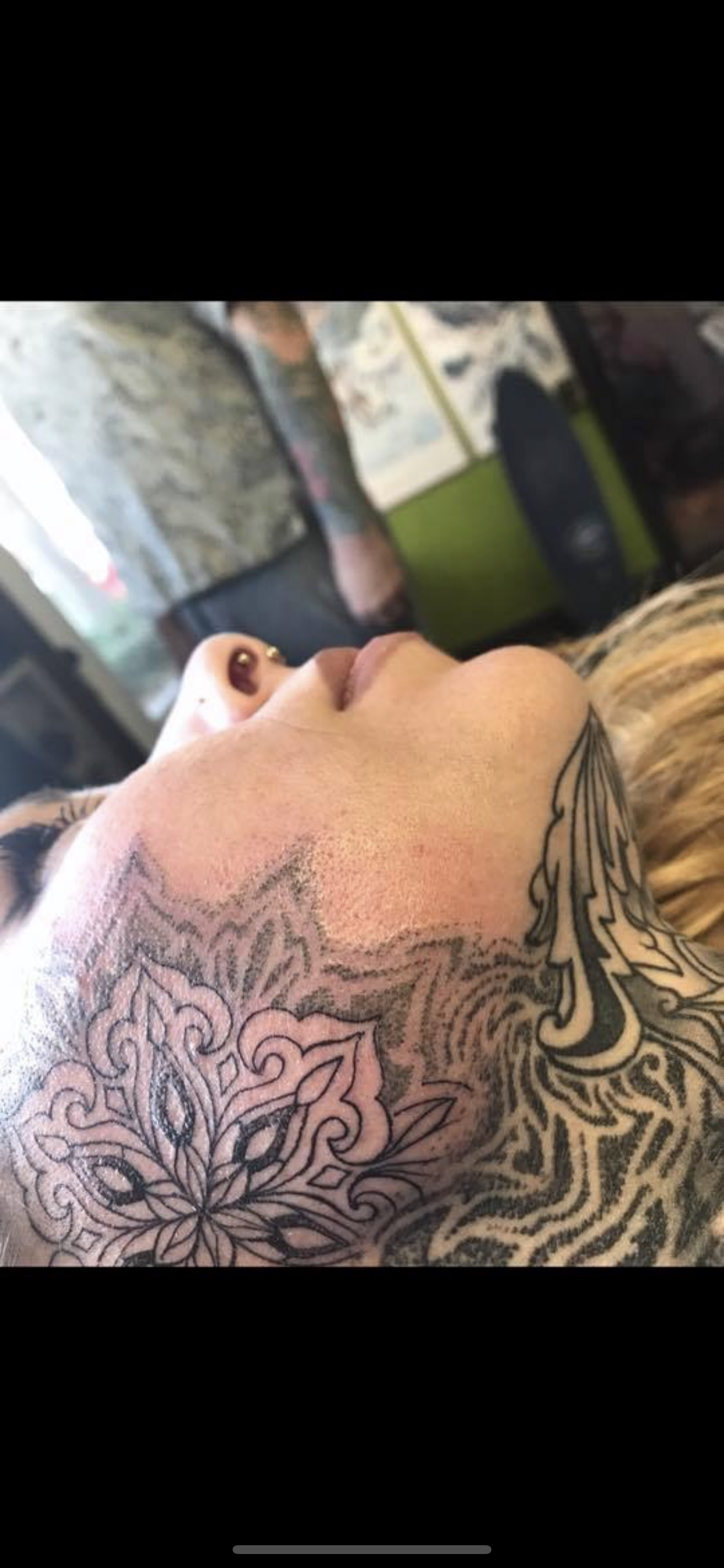 Close-up of a person with facial tattoos, lying down, with a tattooed arm visible. The background shows an indoor setting with a speaker and artwork on the wall.