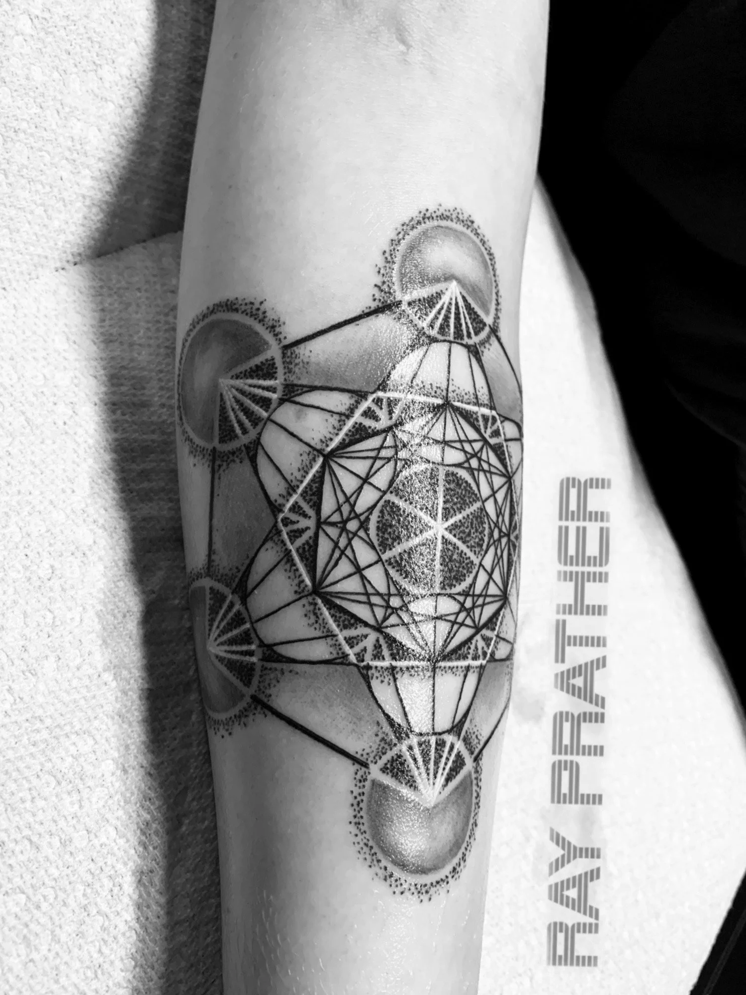 Black and gray geometric tattoo on a person's forearm featuring multiple interconnected shapes including circles, triangles, and a cube, with shading and dot work for depth.