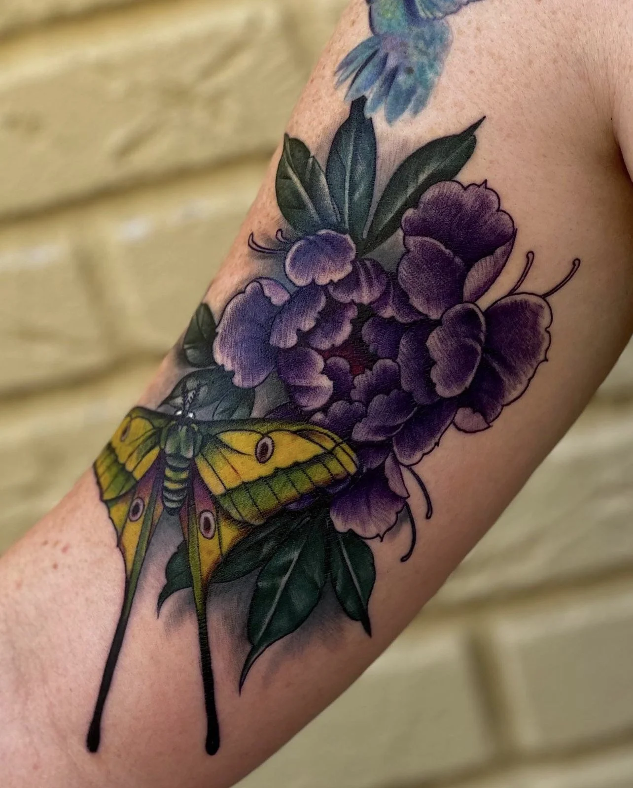 Colorful tattoo of a yellow and green moth resting on purple flowers with green leaves.