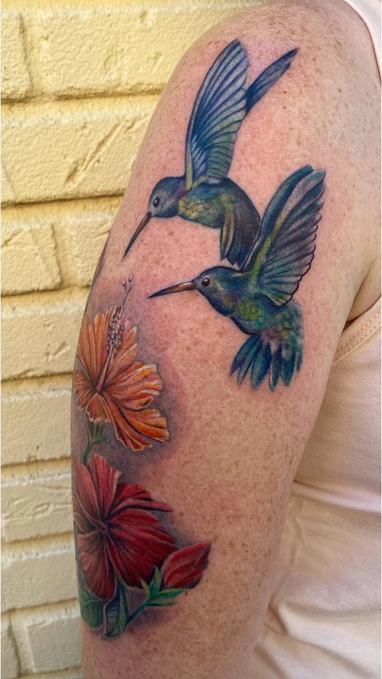 Tattoo of two hummingbirds flying above three orange and red hibiscus flowers on a person's upper arm.