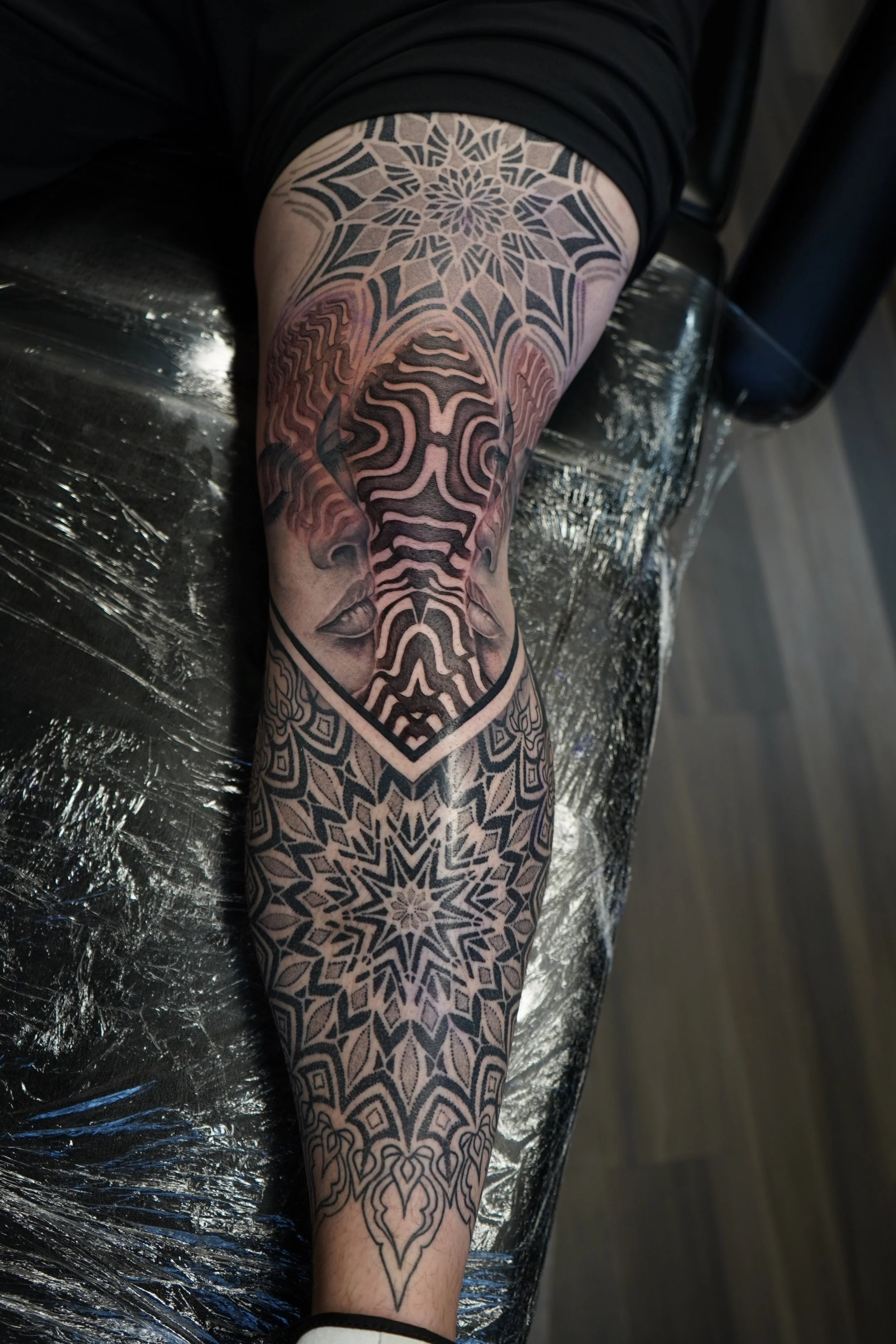 A person's leg with a detailed black and gray tattoo featuring a symmetrical mandala pattern on the lower leg, a stylized face of a woman with closed eyes and flowing hair, and a geometric design at the upper thigh, all outlined with dark lines and s