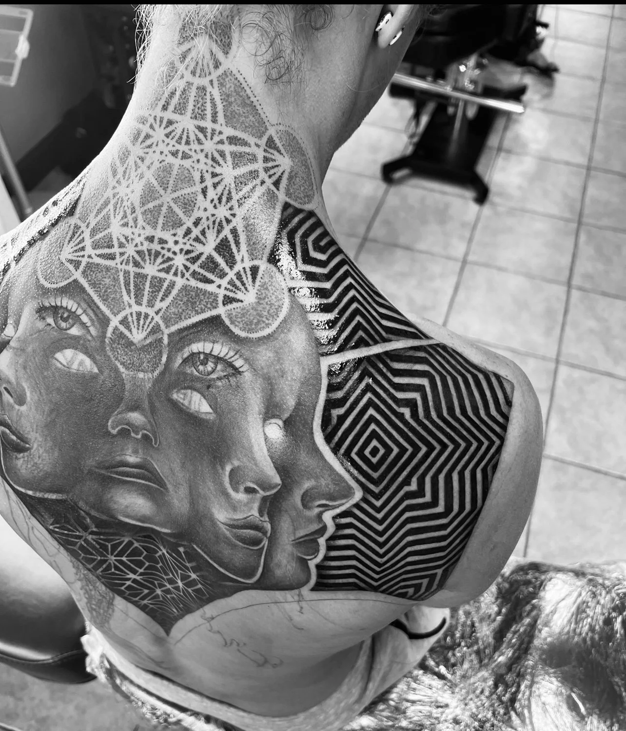Black and gray tattoo on person's upper chest and shoulder featuring a large abstract geometric pattern, a woman's face profile, and three eyes in a surrealistic style.