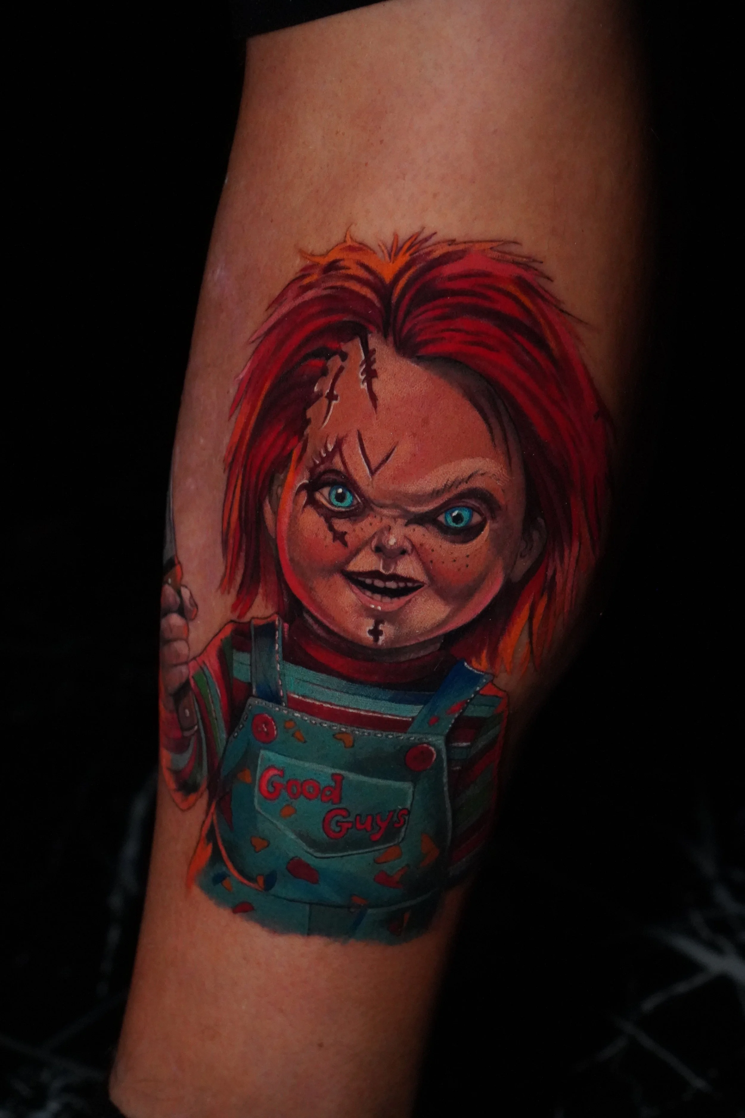 Tattoo of a creepy doll's face with red hair, blue eyes, scars, and a mischievous expression, wearing a striped shirt and a denim overalls with the words 'Good Guys' on the front pocket.