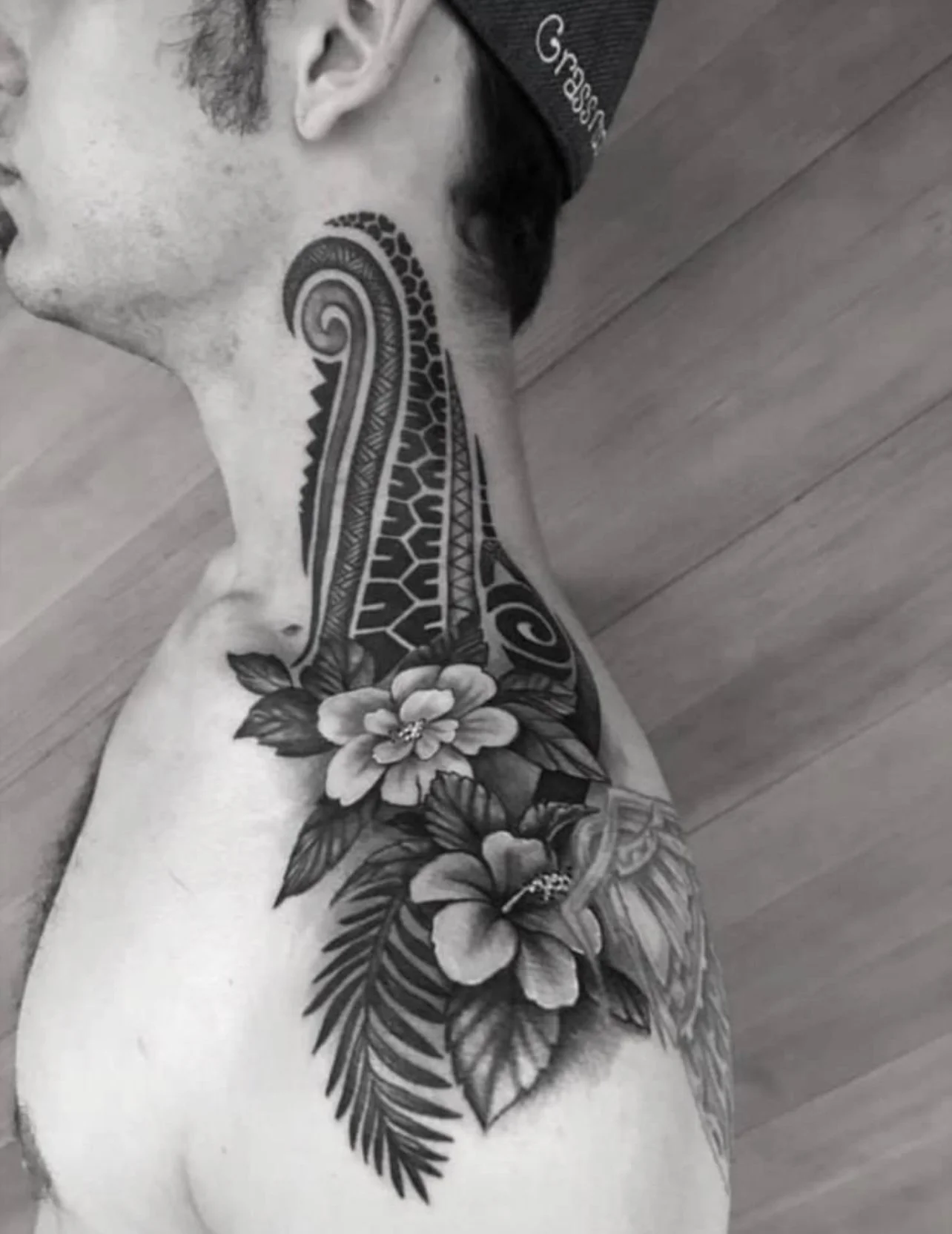 Black and white photo of a man lying on the floor with tattoos on his neck and shoulder, featuring flowers and intricate patterns.