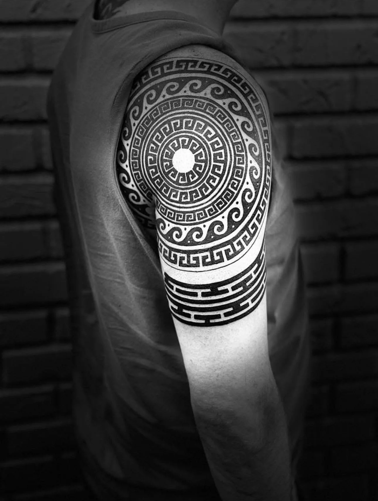 Black and white photo of a person's upper arm with an intricate, geometric tattoo from shoulder to elbow, featuring circular, spiral, and Greek key patterns against a brick wall background.