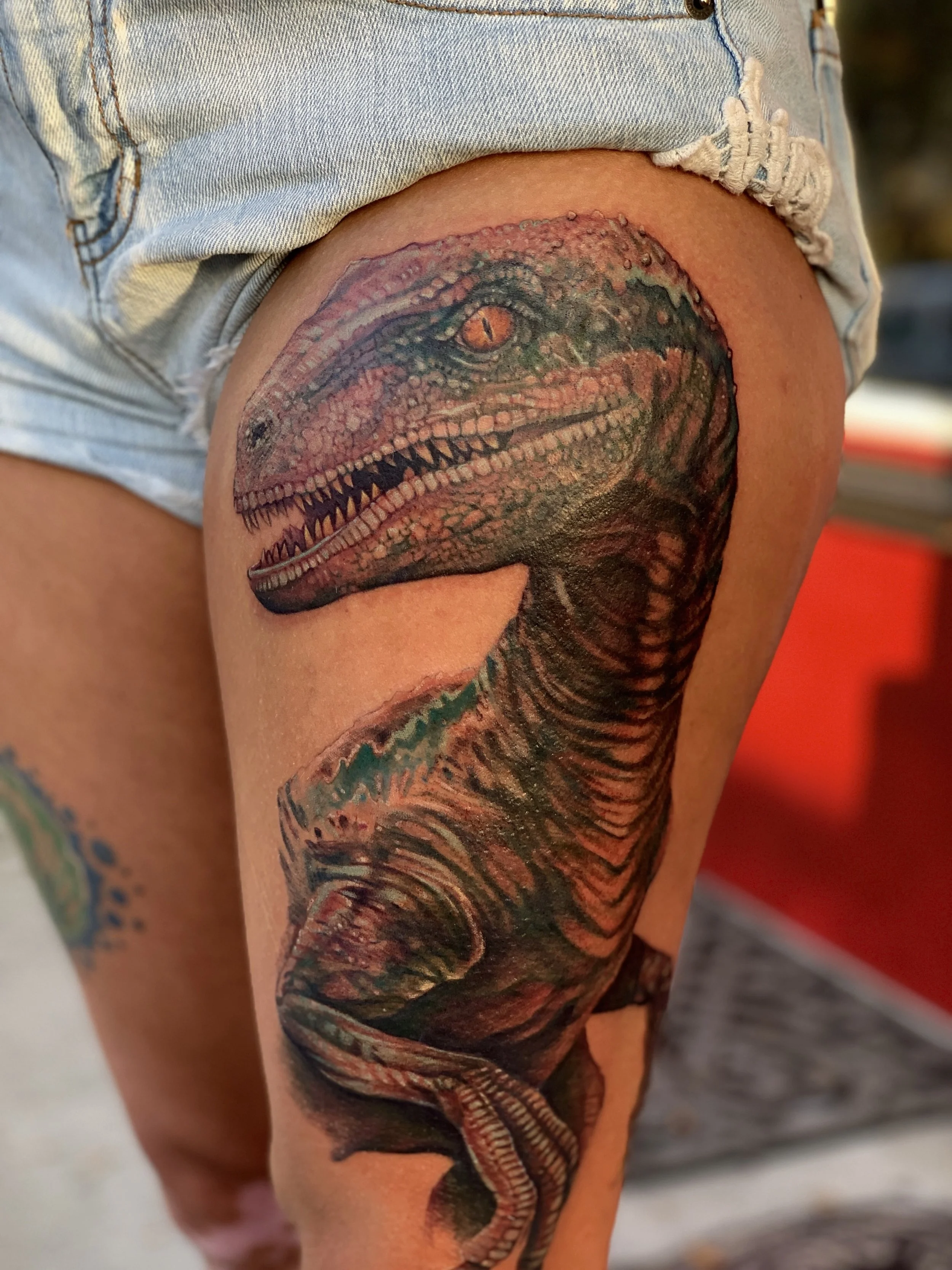 Colorful dinosaur tattoo on person's upper thigh, depicting the head and part of the body, with sharp teeth and detailed scales.