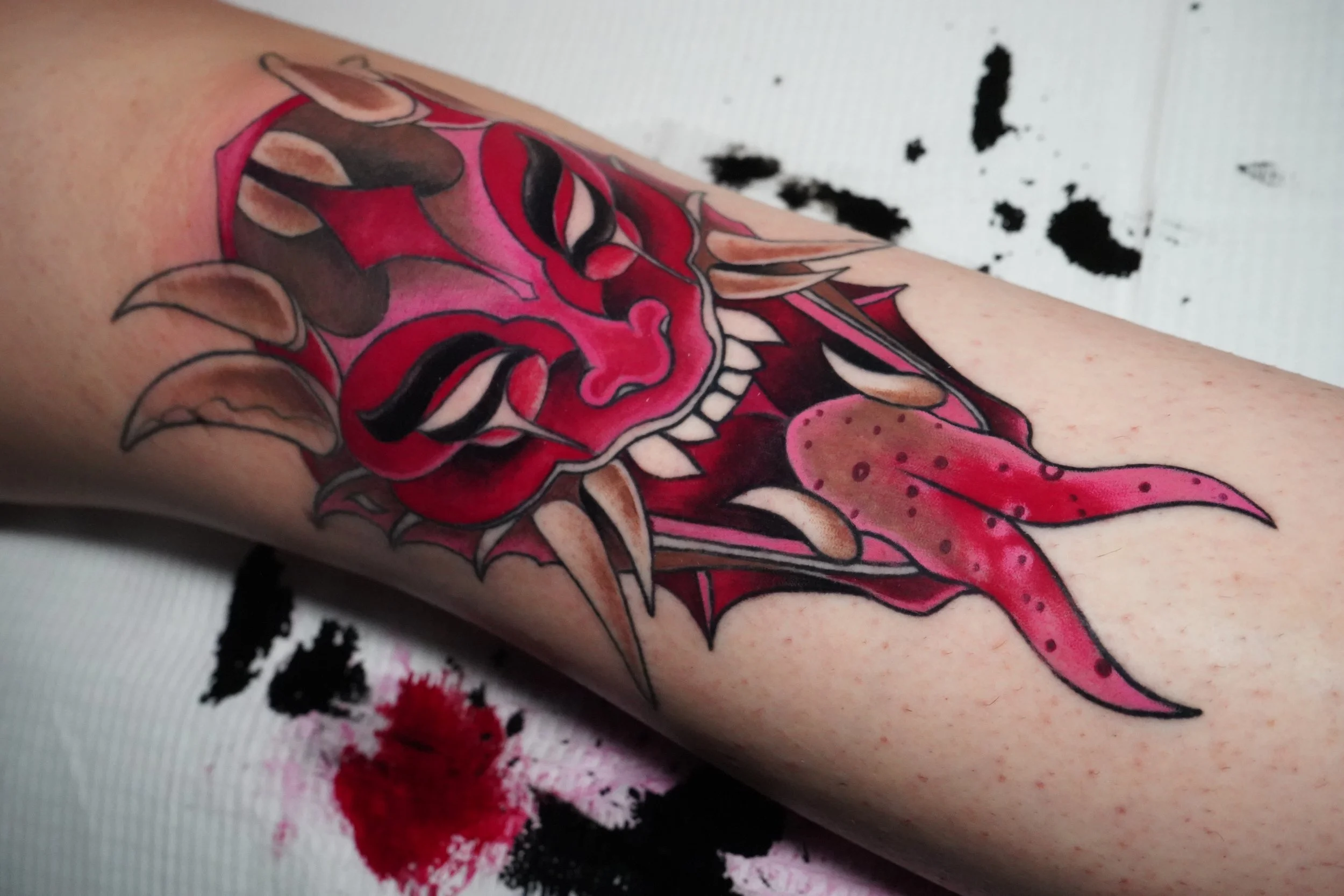 A colorful tattoo on a person's forearm featuring a demonic face with red skin, black and white eyes, and sharp teeth, surrounded by pink and beige flames or tentacles.