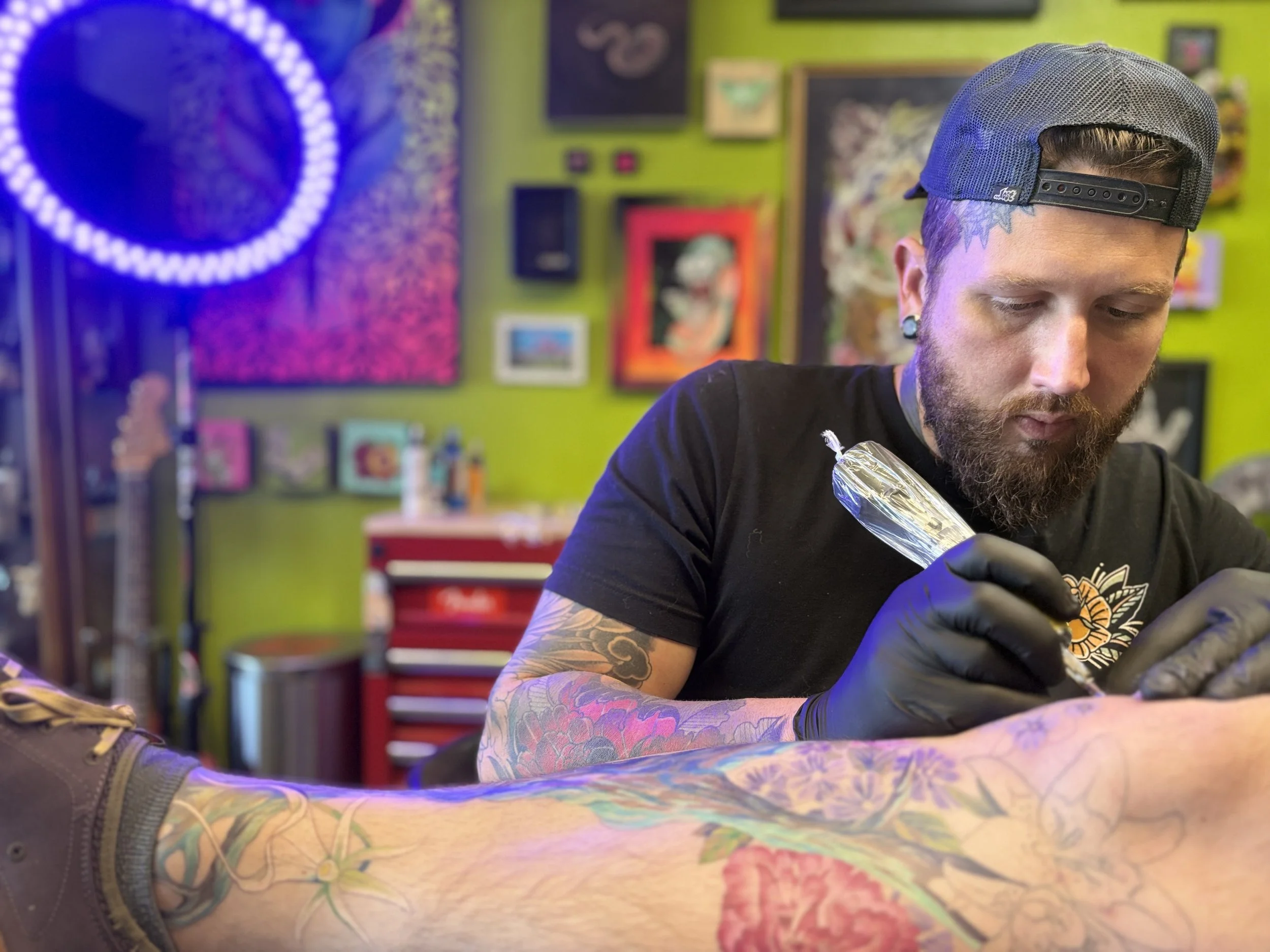 Tattoo artist tattooing a person's leg in an art studio with colorful artwork on the bright green walls.