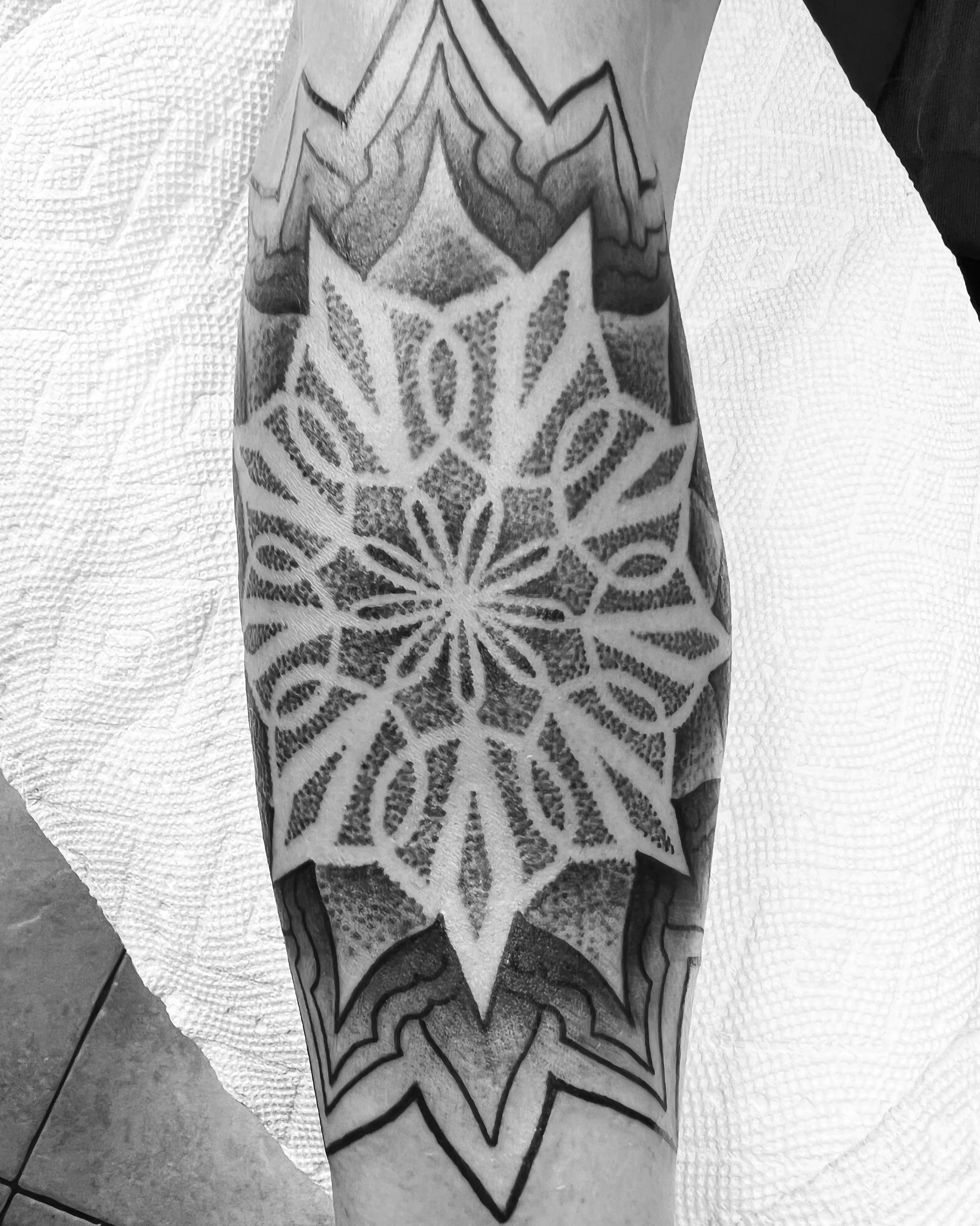 A black and white photo of a detailed geometric tattoo on a person's forearm, featuring a symmetrical starburst pattern with intricate line work and shading.
