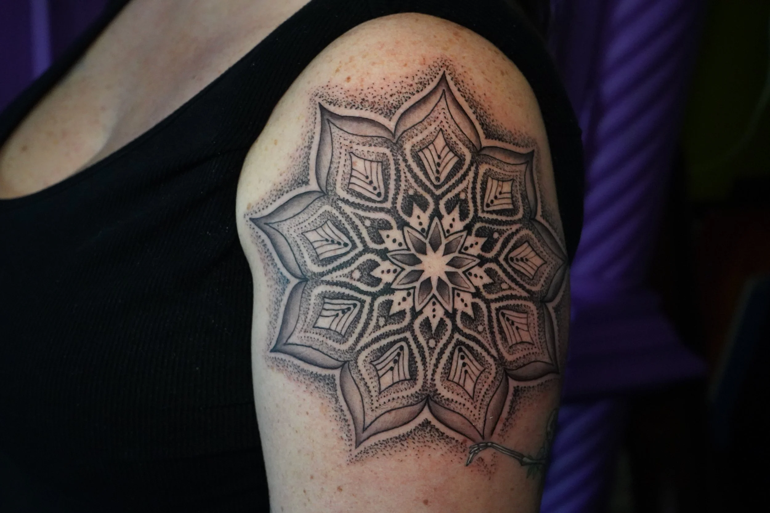 Black and gray geometric floral tattoo on person's upper arm featuring a symmetrical pattern with petal shapes and dotted shading.