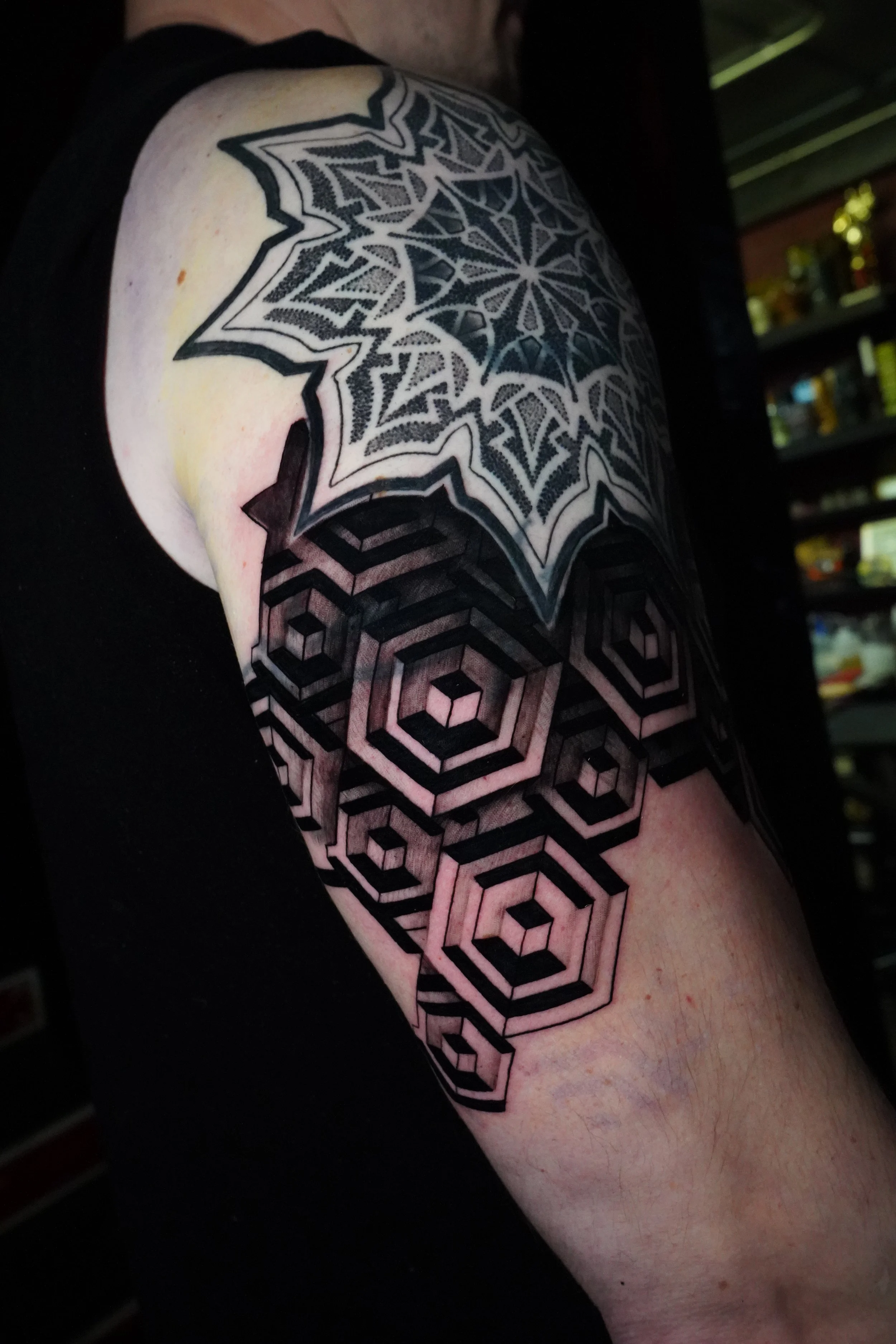 A person's upper arm with a geometric tattoo design featuring black outlines and patterns, including hexagons and a star-shaped pattern.