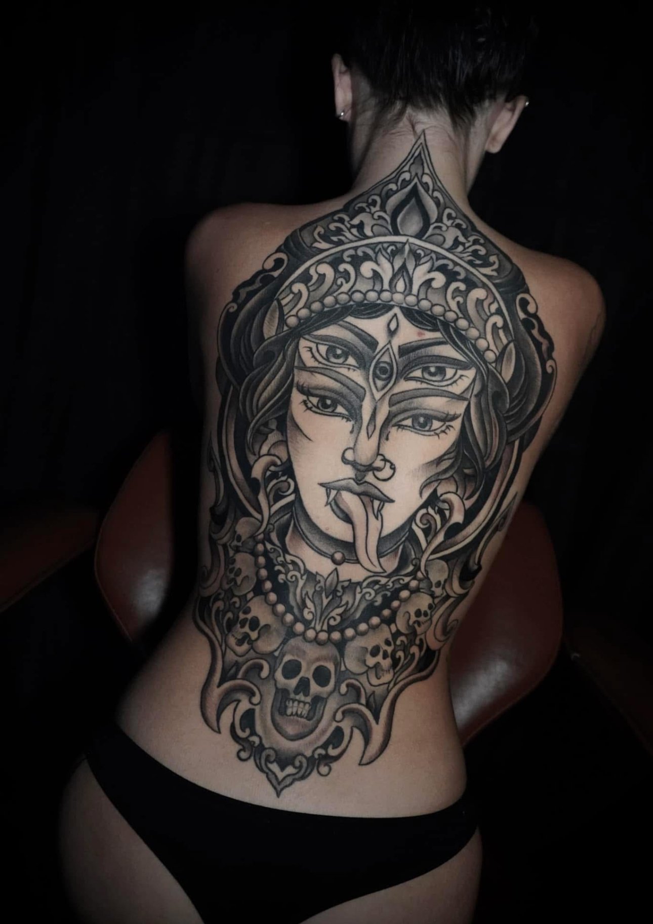 Back tattoo of a multi-eyed woman with a crown, snake, and skull in black and gray ink.