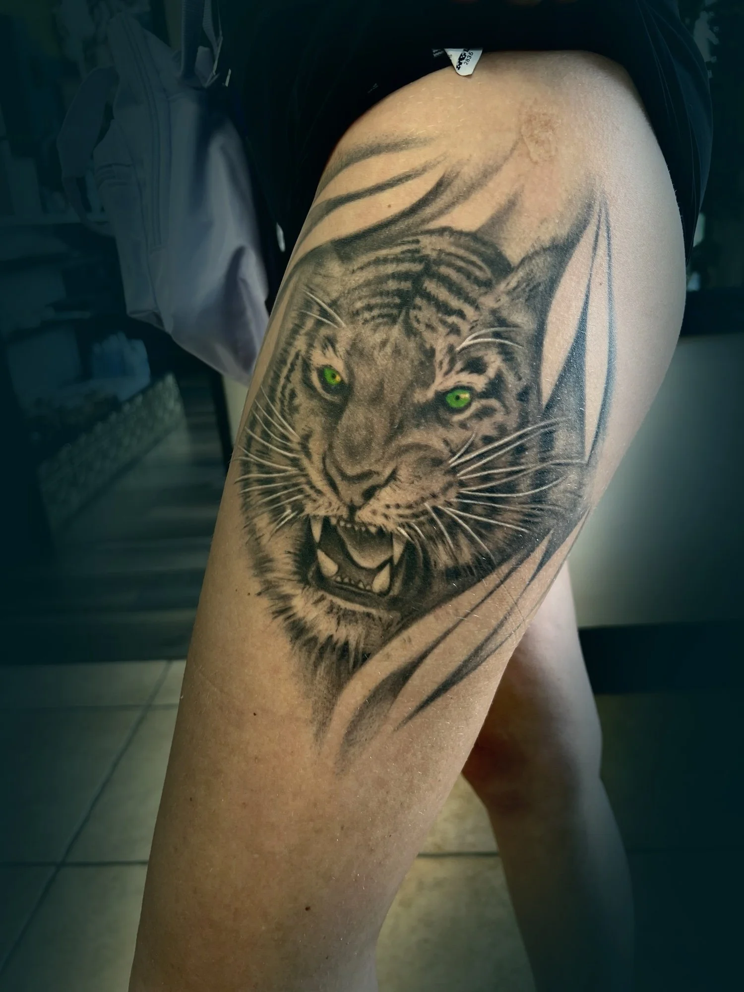 Tattoo of a roaring tiger with green eyes on a person's thigh.