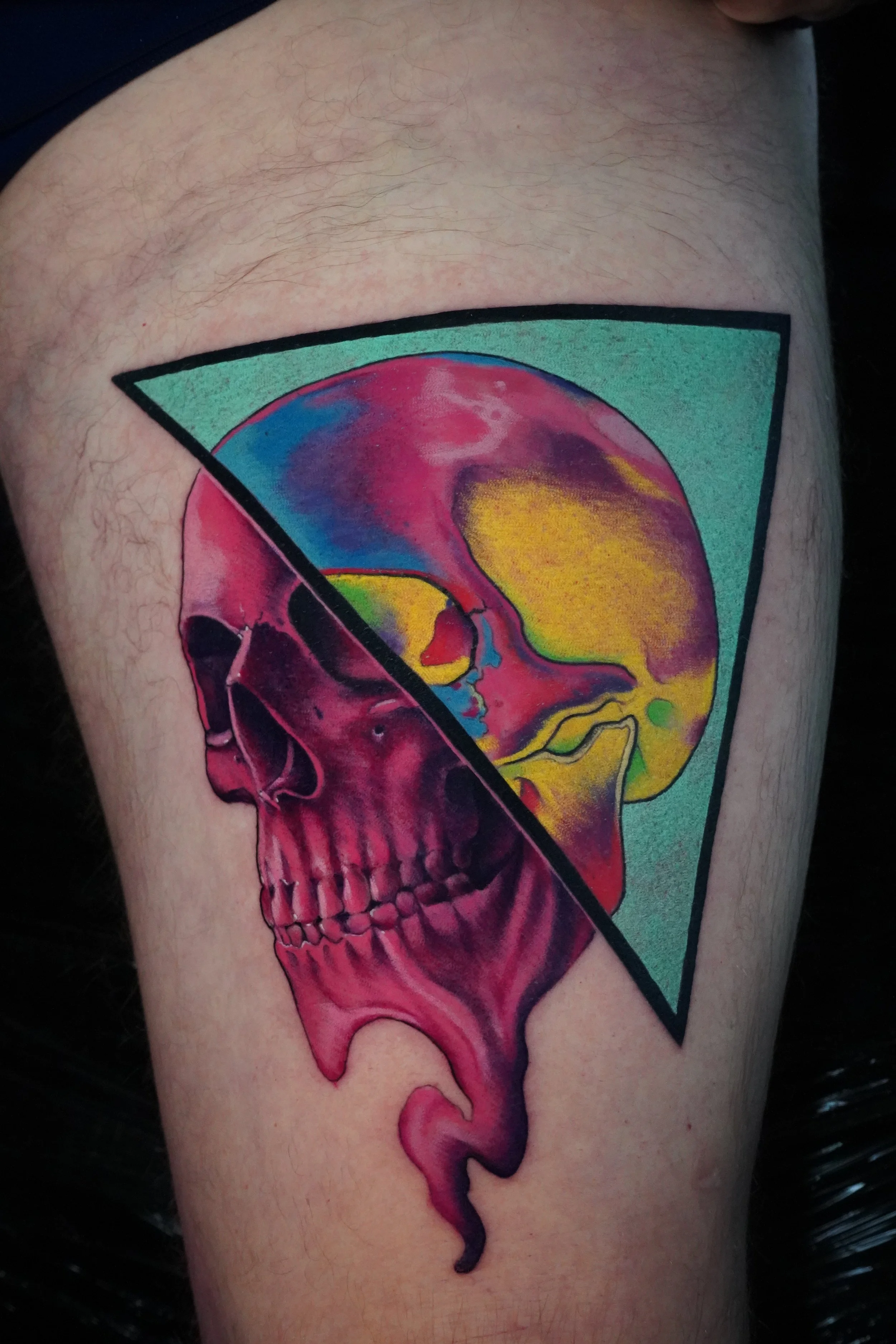 Colorful skull tattoo partially inside a green triangle outline on a person's thigh.