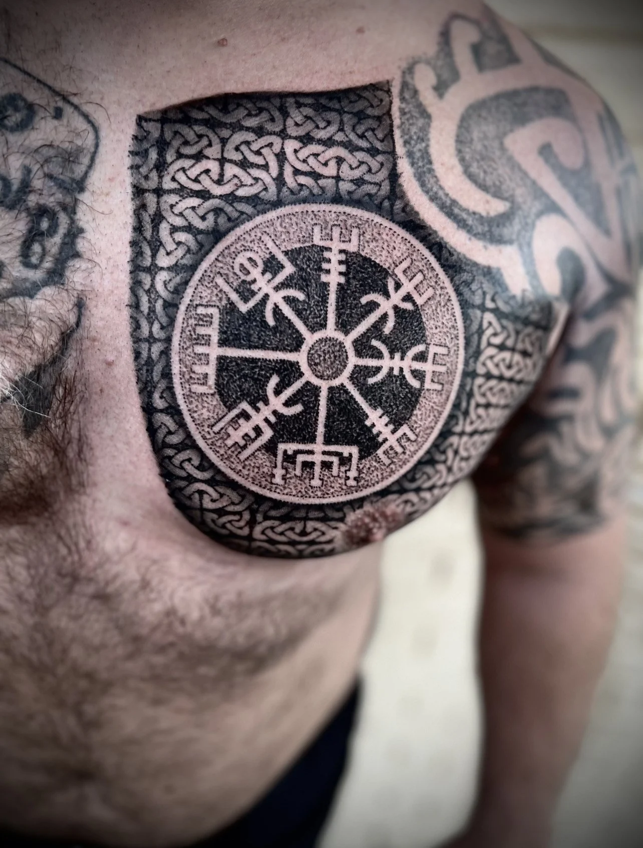 Close-up of a person's chest with a large, intricate black and gray tattoo featuring a Norse symbolism or runic design.